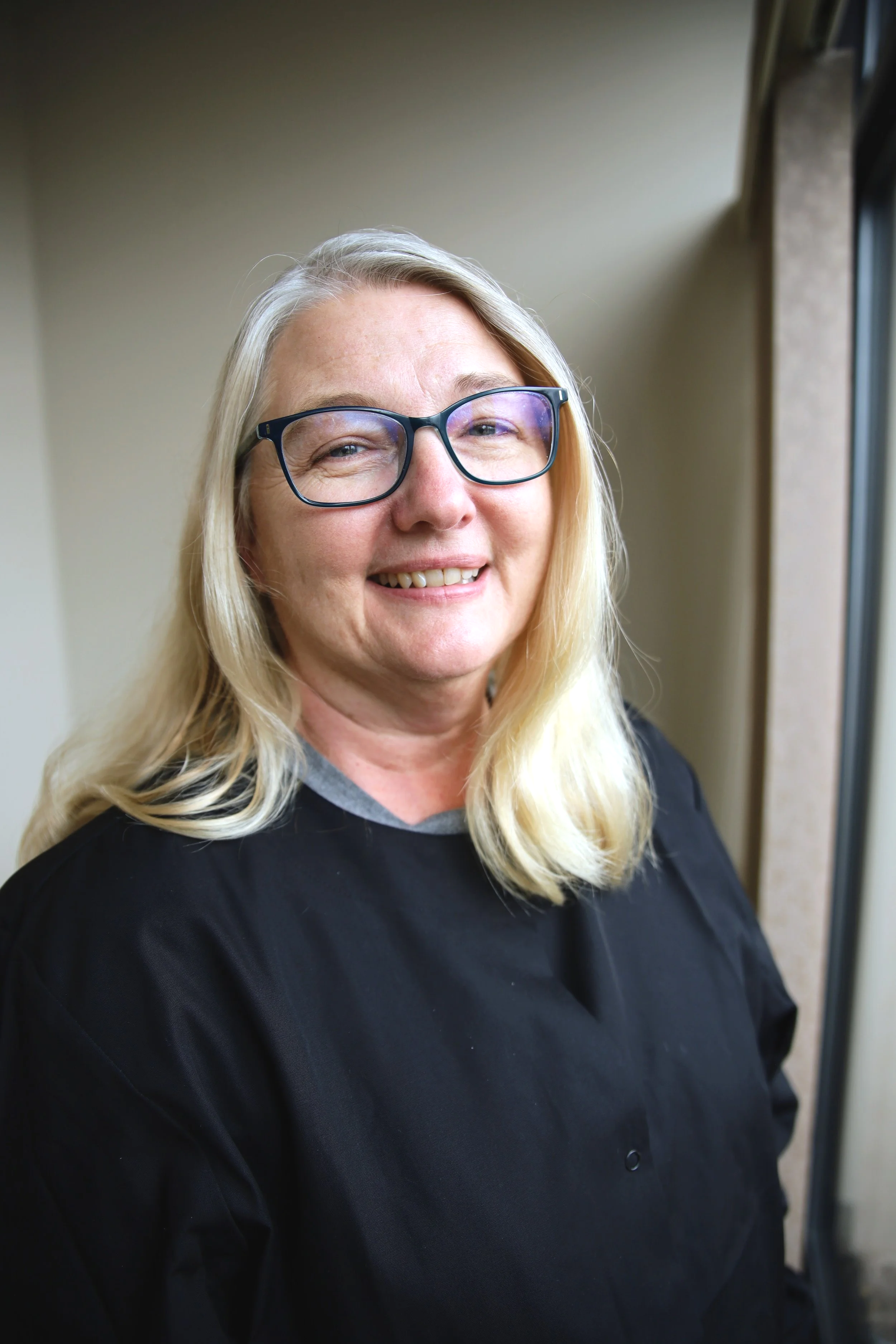 A smiling woman with blonde hair and glasses wearing a black shirt, standing indoors near a window.