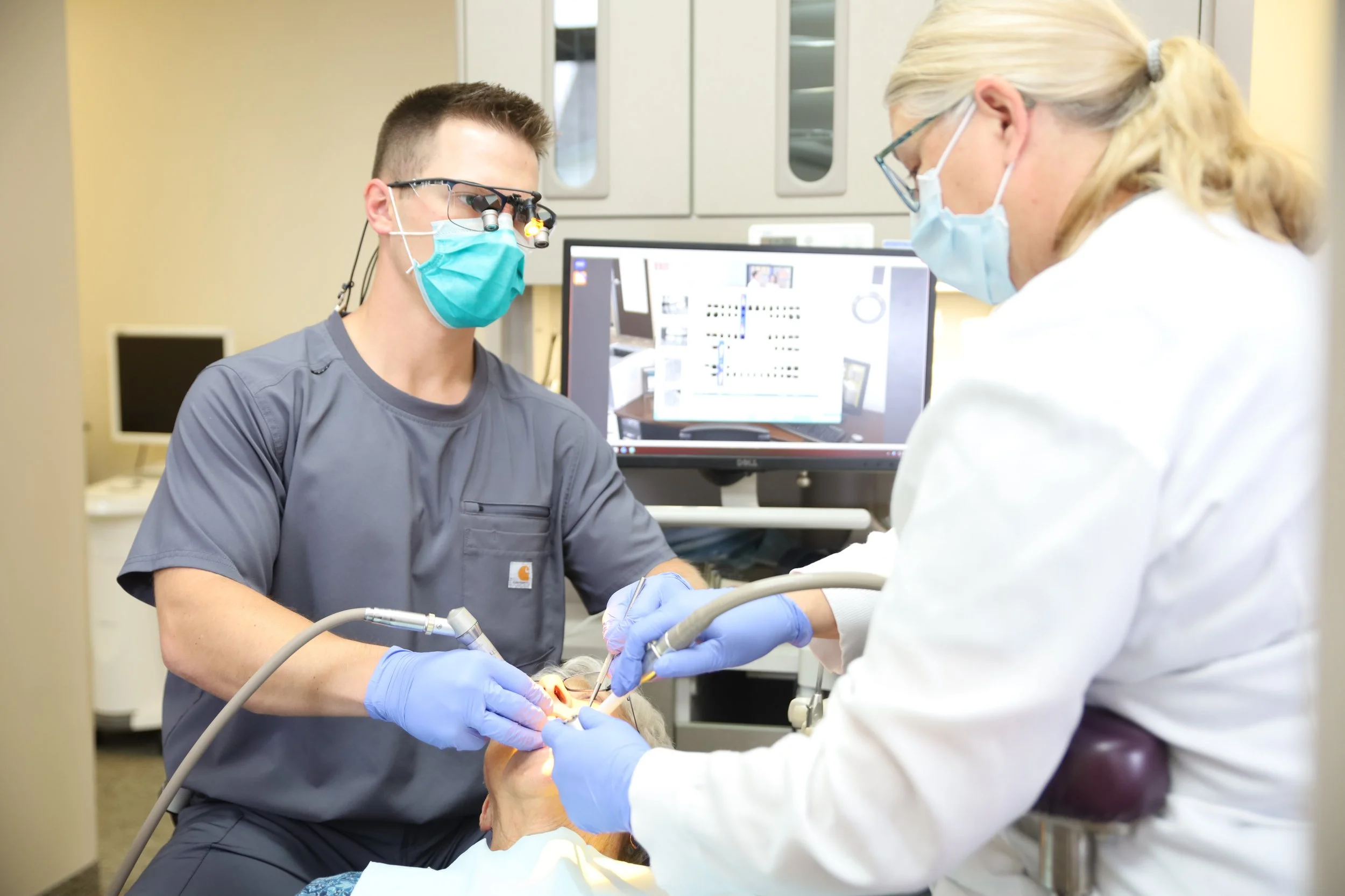 A male dentist wearing scrubs, a surgical mask, and magnifying glasses with a headlamp, performs dental work on a patient in a dental clinic, with a female dental assistant wearing a face mask and gloves assisting. A computer monitor in the background displays dental images.