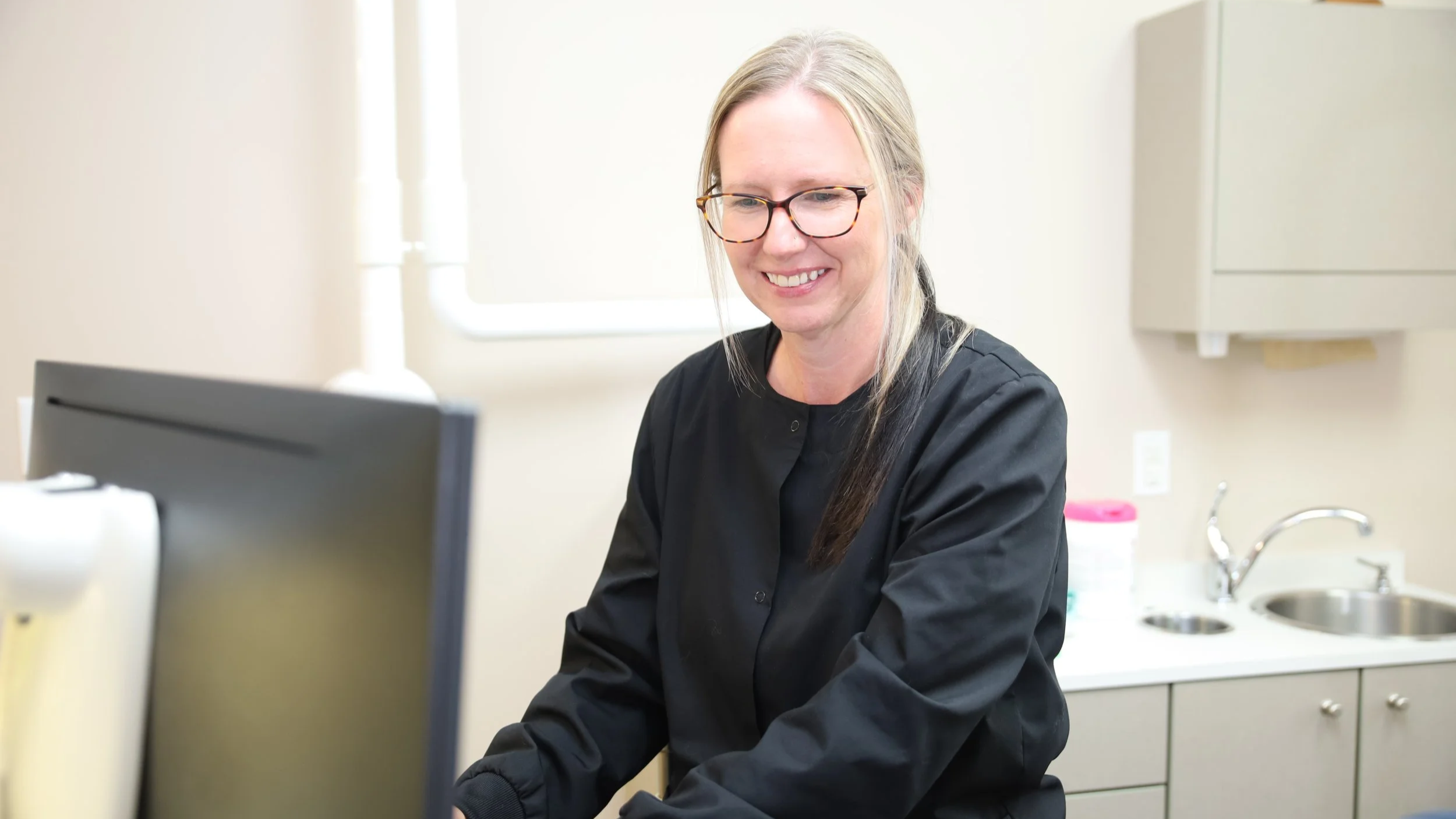 A woman with glasses and long blonde hair smiling while working at a computer in a medical or laboratory setting.