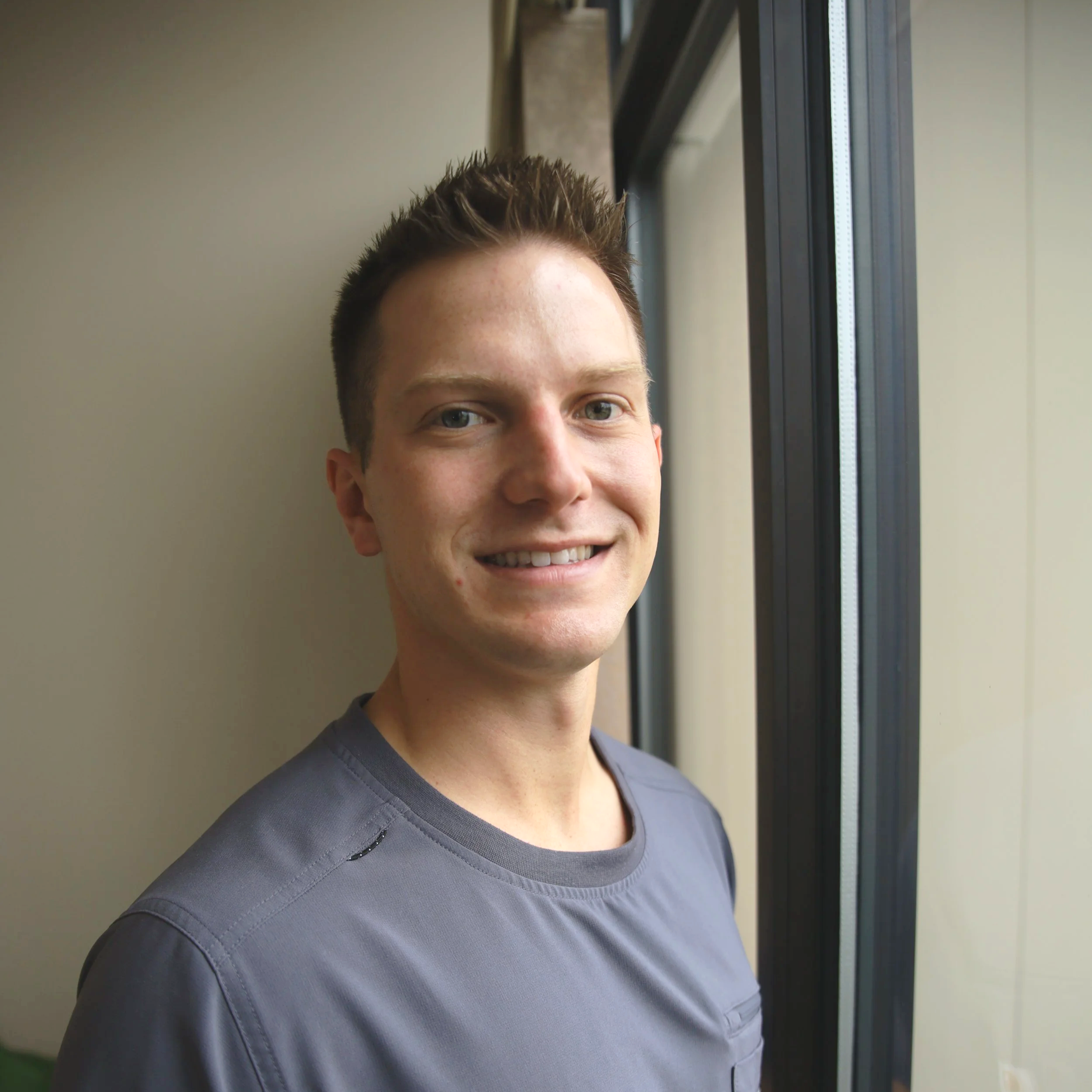 Close-up of a young man with short spiky brown hair and blue eyes, smiling slightly, standing near a window with natural light illuminating his face, wearing a dark gray shirt.