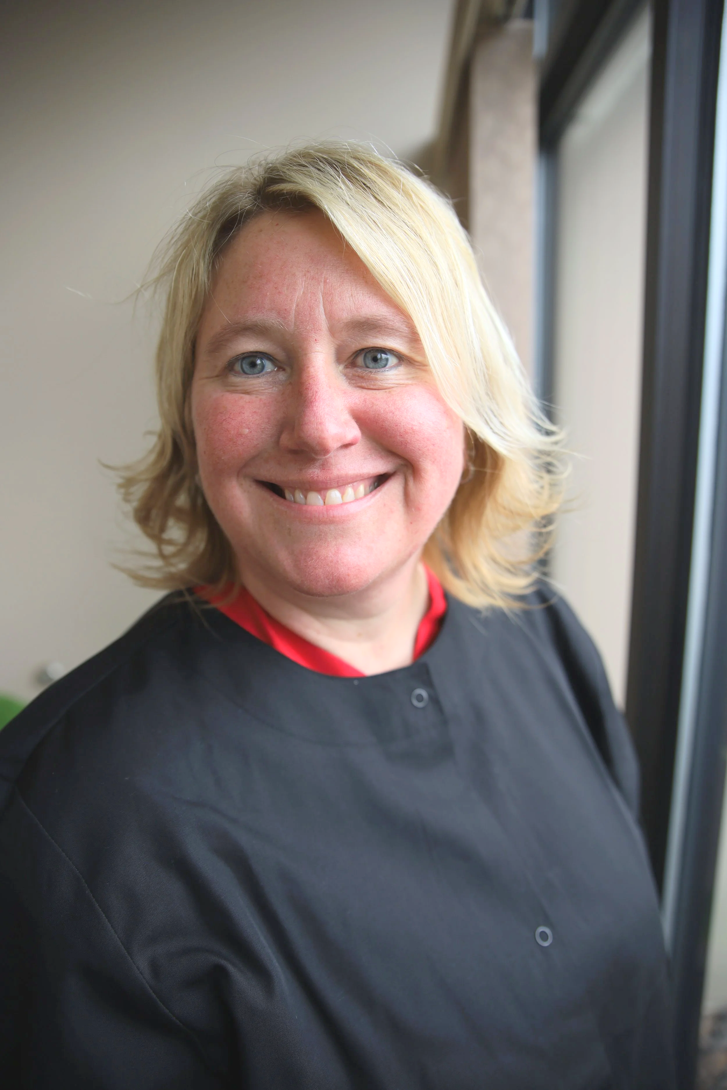 Close-up of a smiling woman in a black medical gown standing near a window.