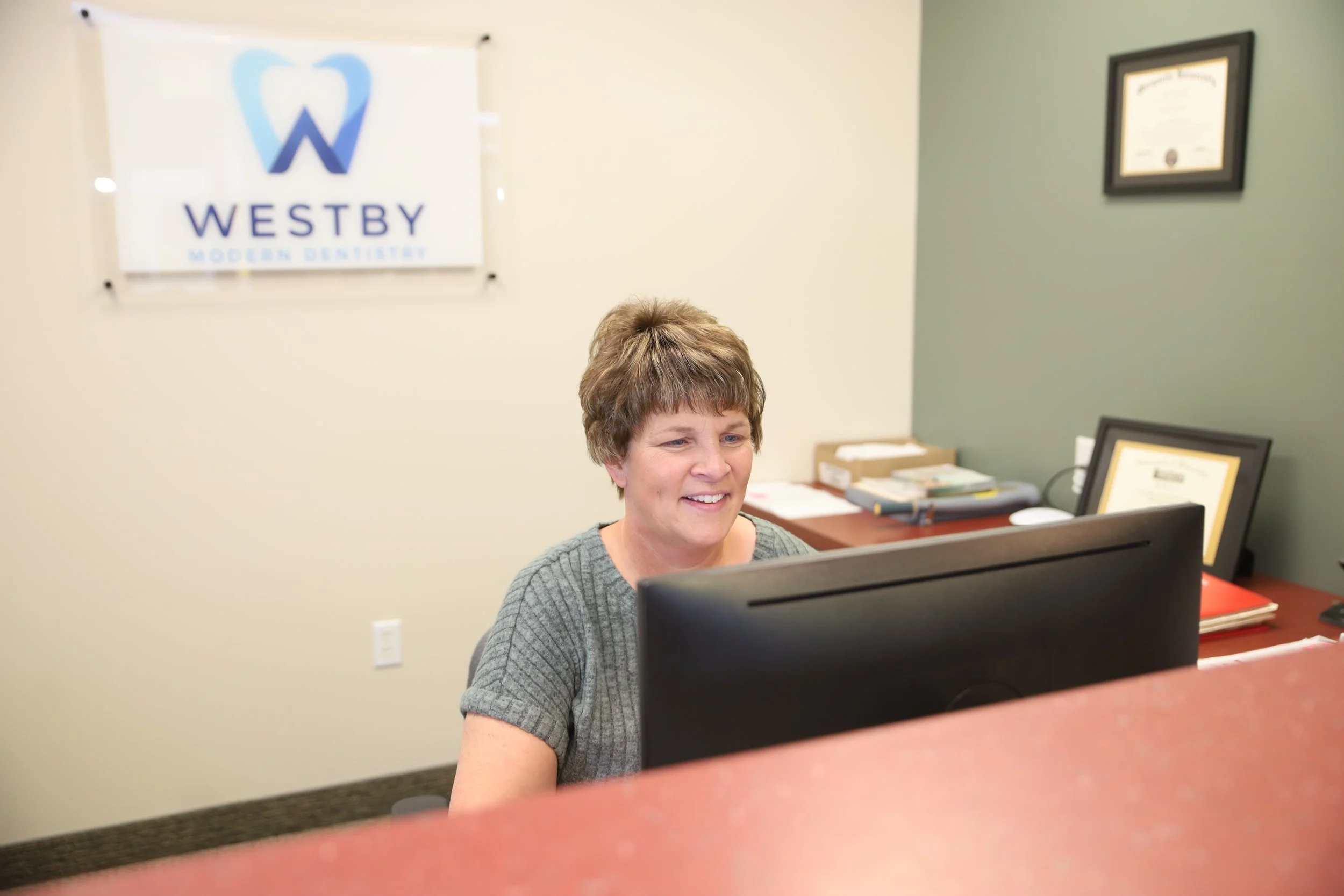 A woman with short hair sitting at a desk in an office, looking at a computer monitor with a smile. Behind her, a sign reads "WESTBY MODERN DENTISTRY" accompanied by a logo. There are framed certificates on the wall and files on the desk.