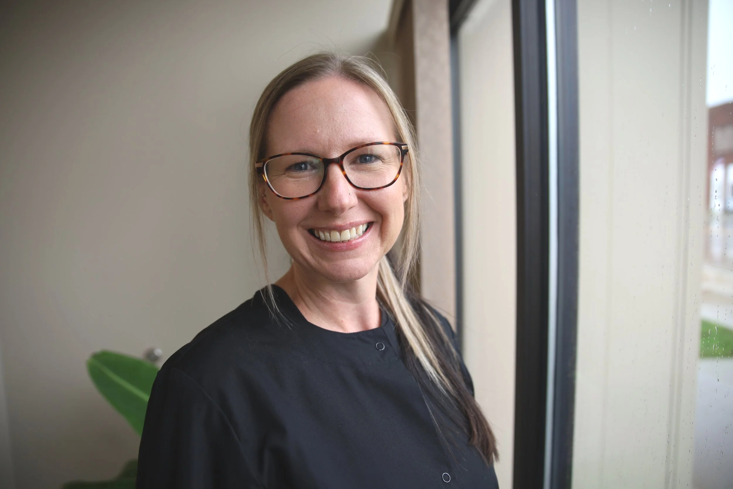 A smiling woman with blonde hair and glasses standing by a window.