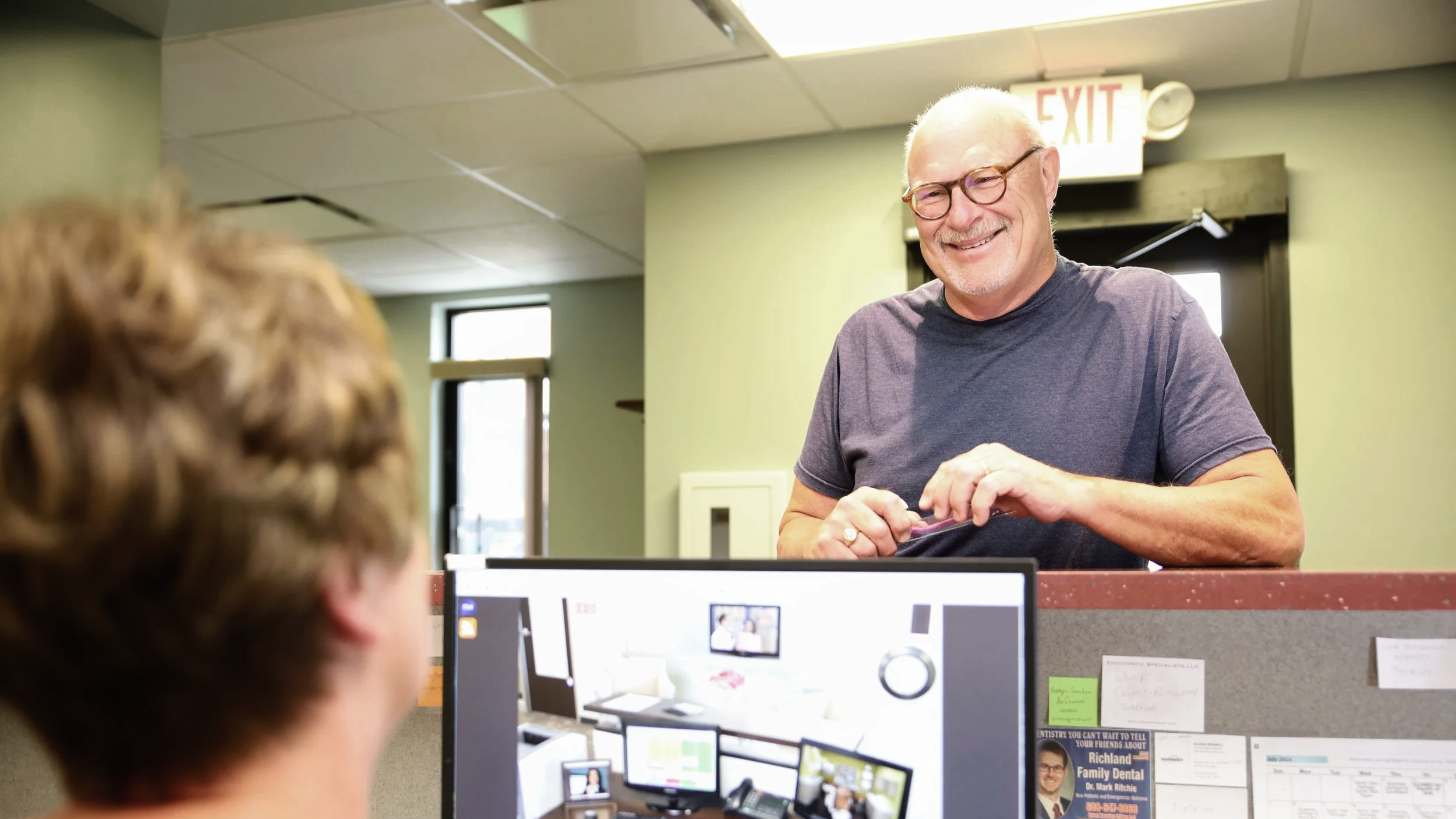 A smiling older man with glasses and a beard, wearing a dark t-shirt, standing at a reception desk and speaking to a person behind a computer in an indoor office or clinic setting.