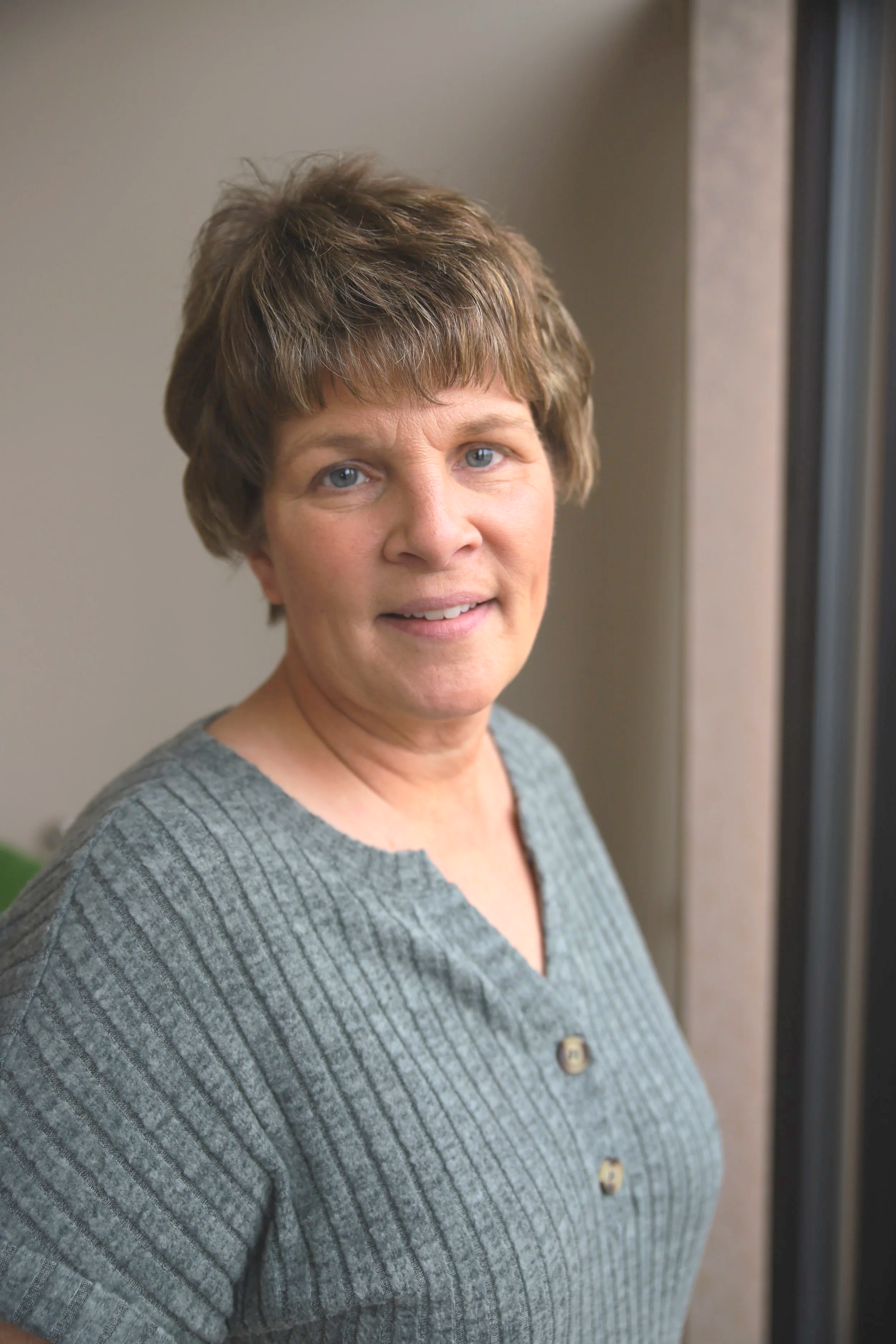 A close-up of a middle-aged woman with short brown hair, light blue eyes, and a slight smile, wearing a gray ribbed sweater with buttons on the front, standing indoors near a window or glass door.