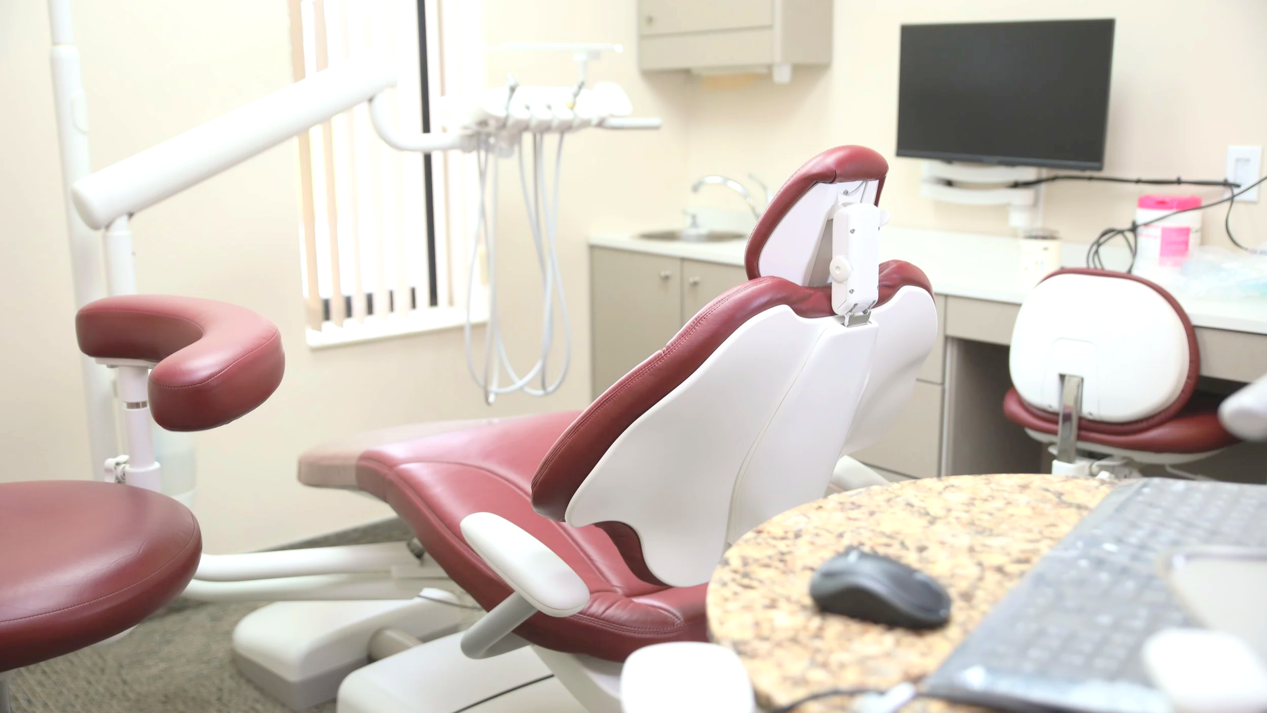 Dental office with a reclining dental chair, a stool, a countertop, and a mounted TV in the background.