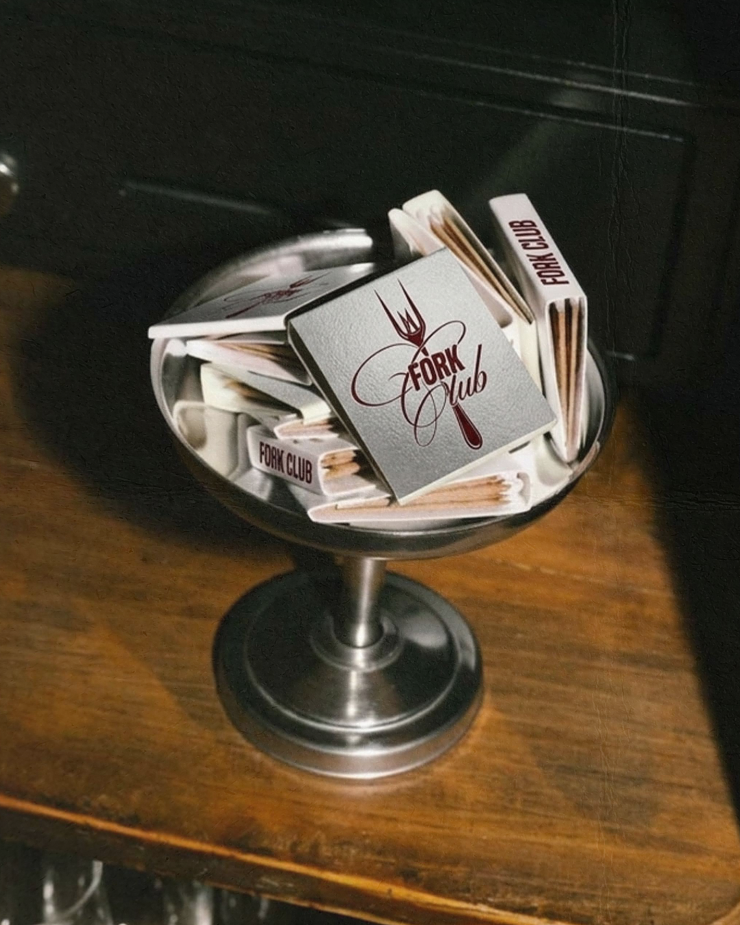 A metal bowl filled with cocktail napkins and matchbooks with the 'Fork Club' logo, placed on a wooden surface.