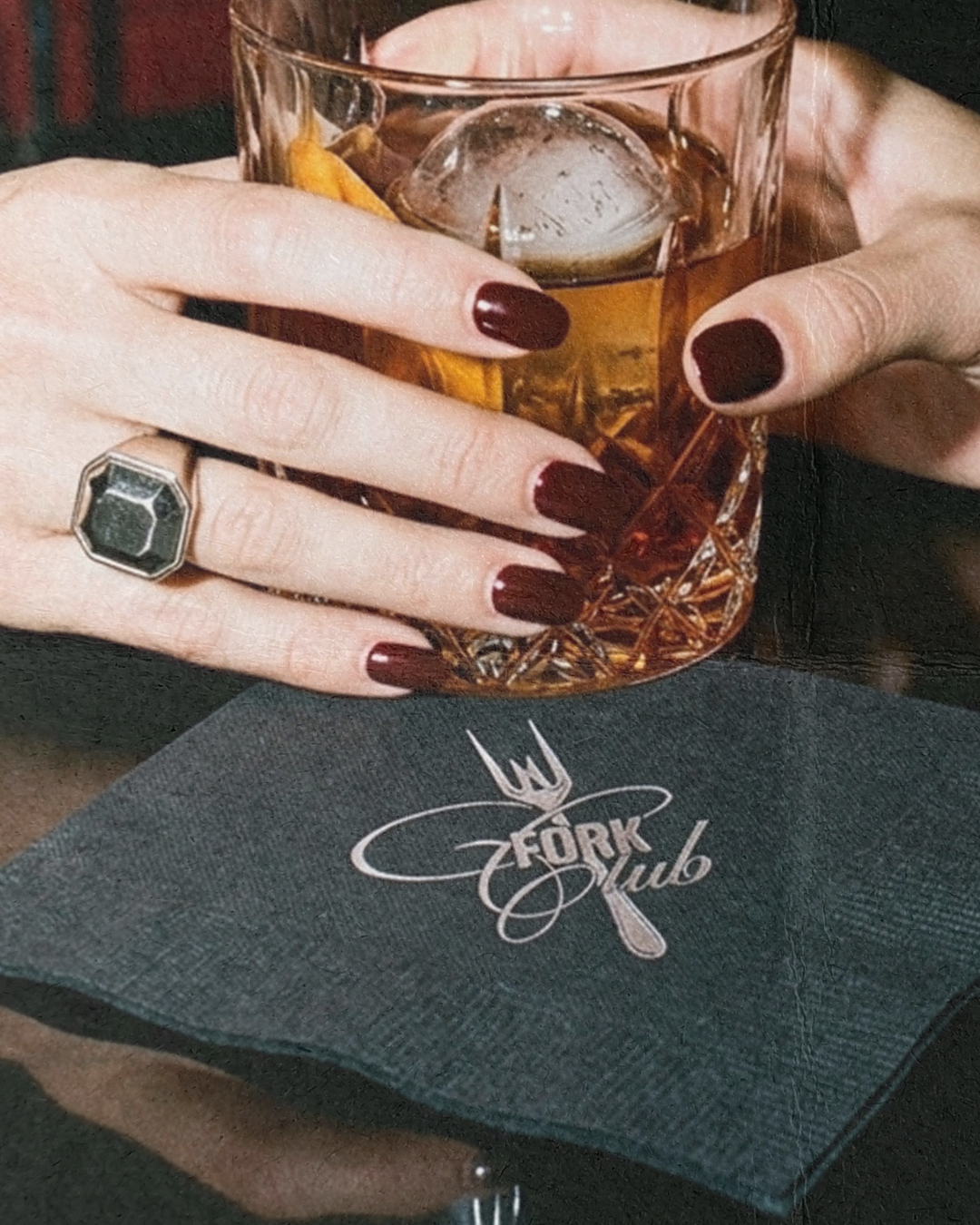 Close-up of a person's hands holding an iced drink in a glass with a textured pattern, on a table with a Fork Club paper napkin, with dark red nail polish and a large geometric ring.