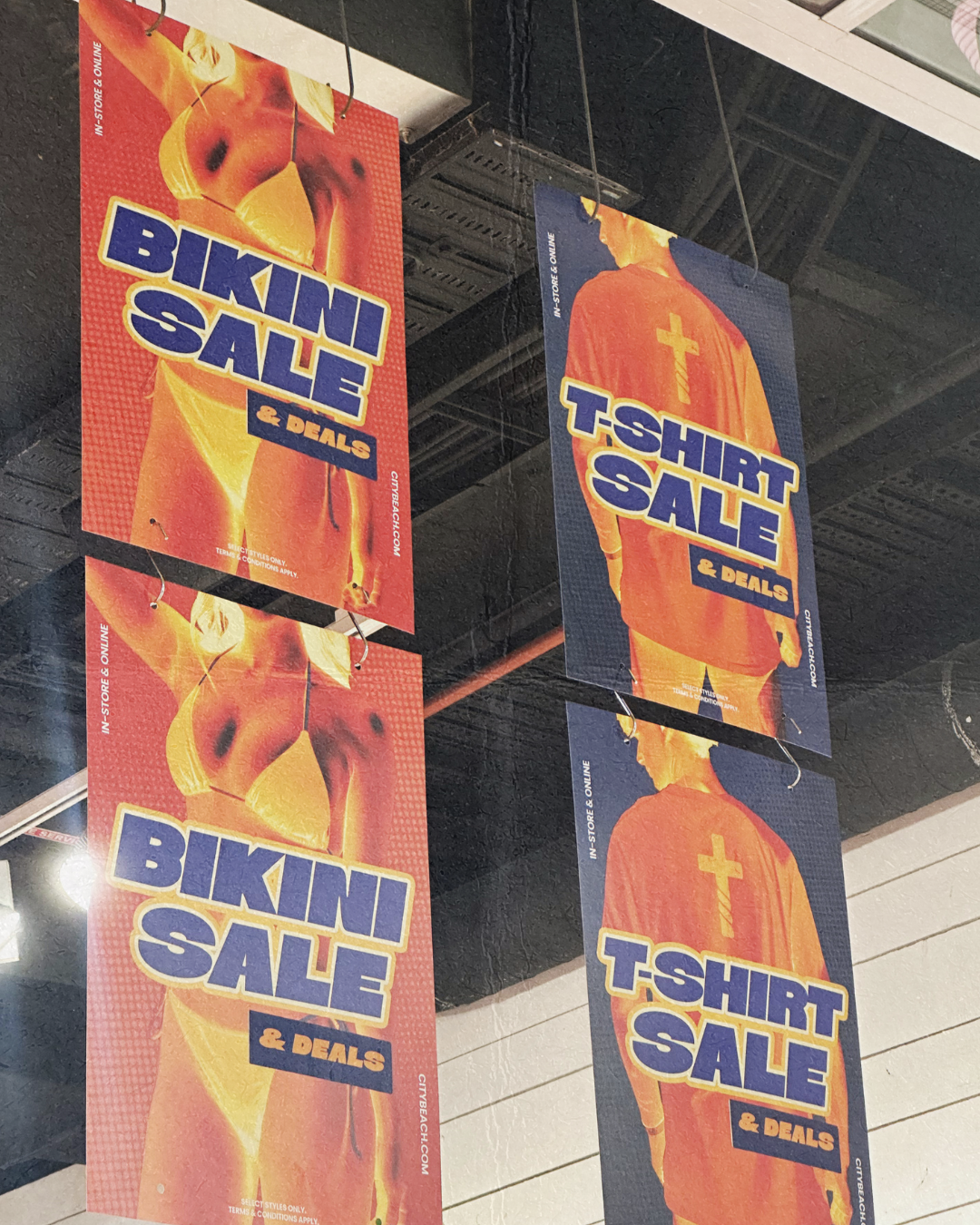 Two vertical banners advertising a bikini sale and a T-shirt sale, hanging from the ceiling. One banner shows a person in a bikini with the text 'BIKINI SALE & DEALS' and the other shows a person from behind wearing a T-shirt with the text 'T-SHIRT S