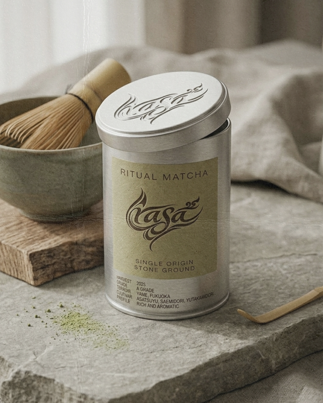 A tin of Casa Ritual Matcha stone ground matcha powder on a gray surface, with a small pile of matcha powder, a wooden spoon, and a bowl with a bamboo whisk in the background.
