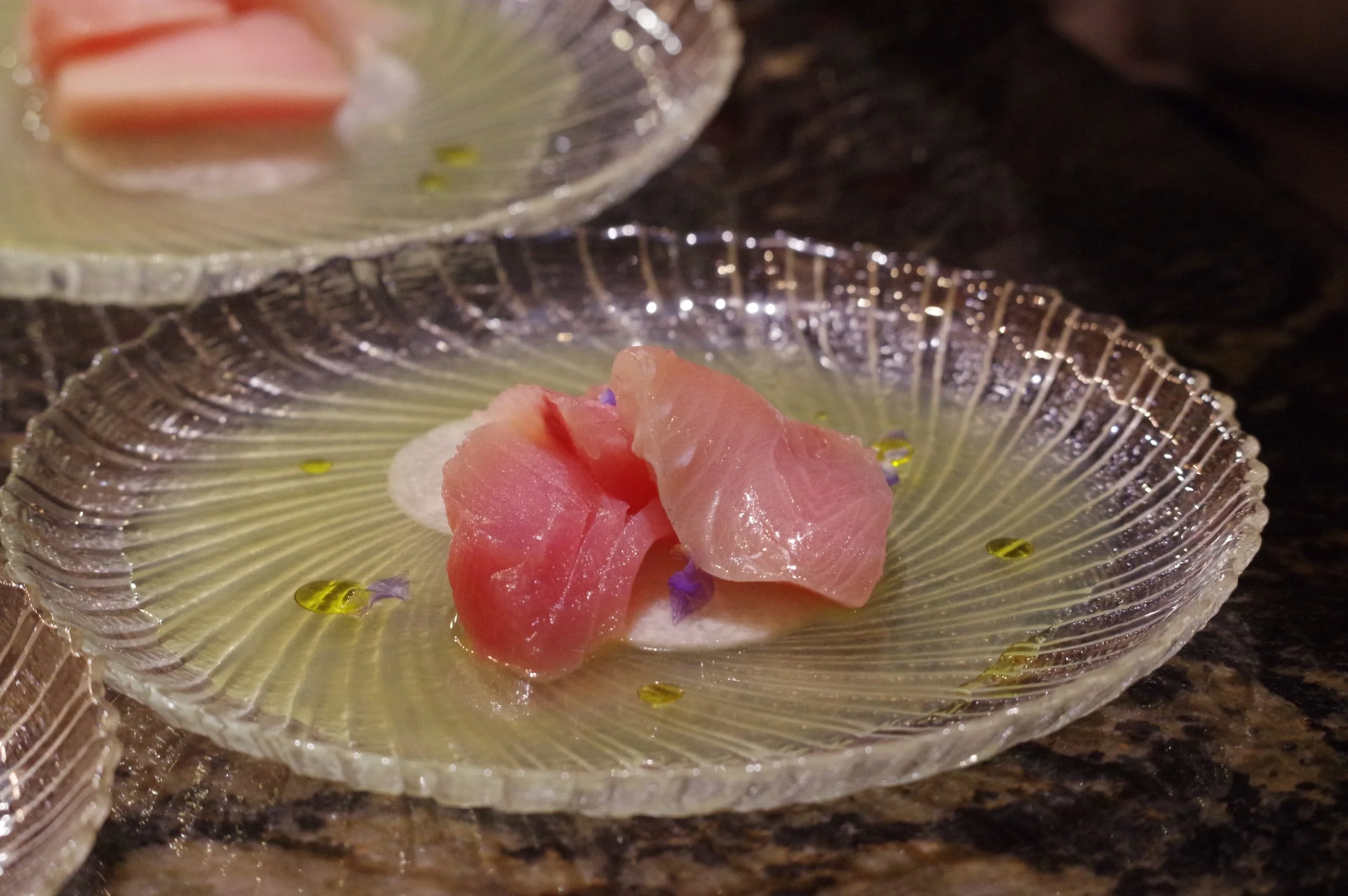Close-up of a glass dish with a pink slice of sashimi garnished with purple edible flowers and tiny yellow and purple beads on a dark surface.