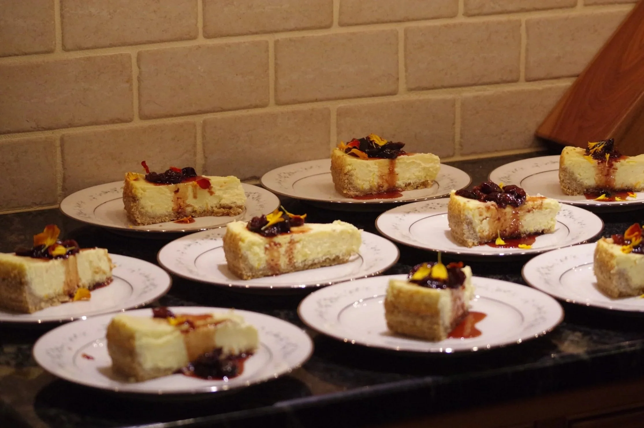Several slices of cheesecake on white plates, garnished with fruit topping and edible flower petals, arranged on a dark surface with a brick wall background.