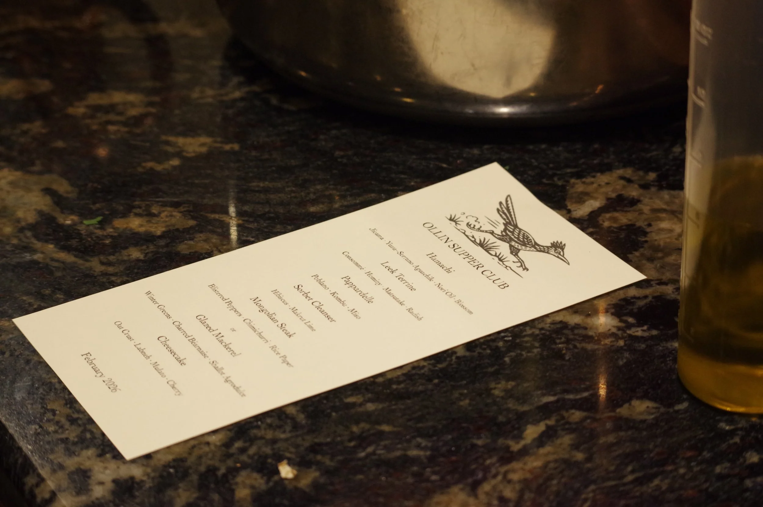 A menu from the HuNachi SUPPER CLUB with details of various food options, placed on a dark, marbled countertop, with a metallic container partially visible at the top and a bottle on the right side.