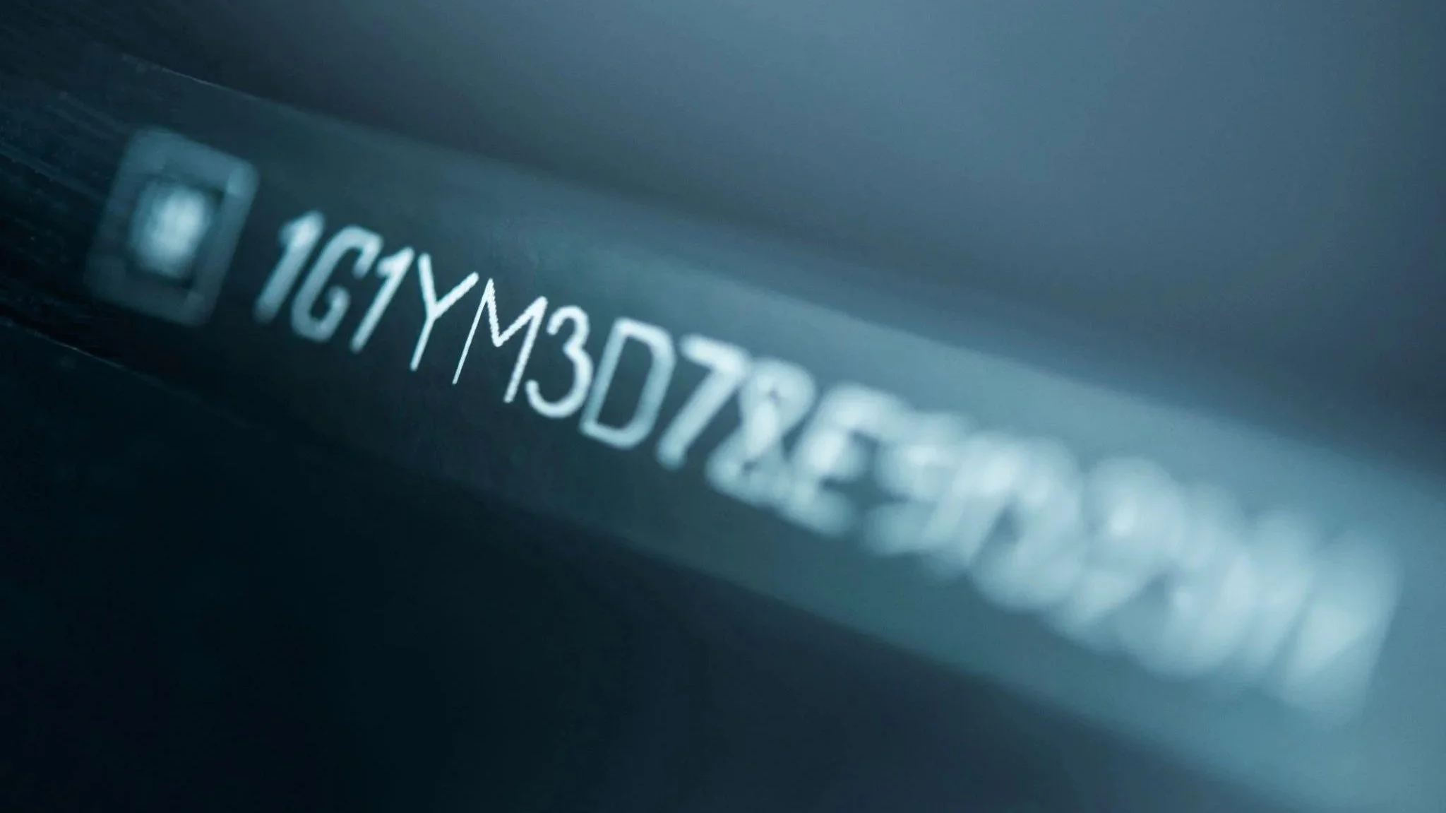 Blurred close-up of a vehicle identification number (VIN) stamped on a metal surface, with characters partially visible.