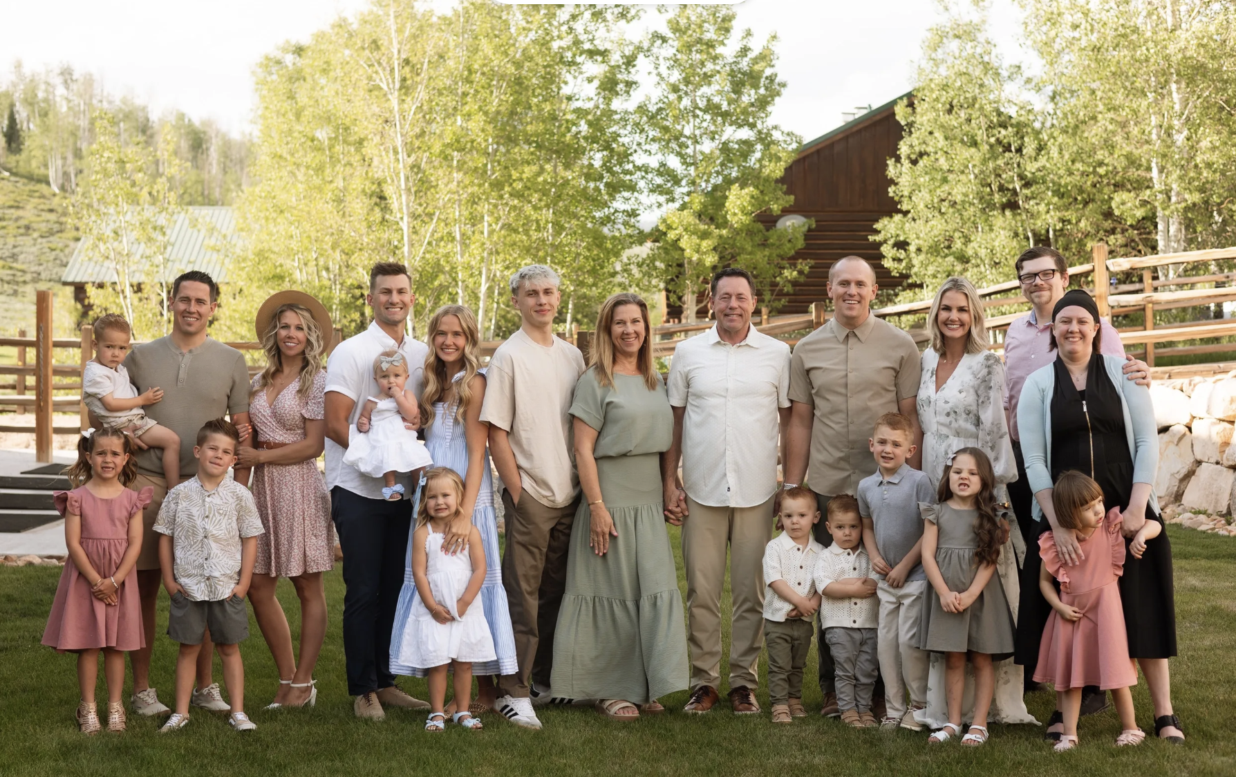 Family photo outdoors with adults and children standing on grass, smiling, with trees and a wooden building in the background.
