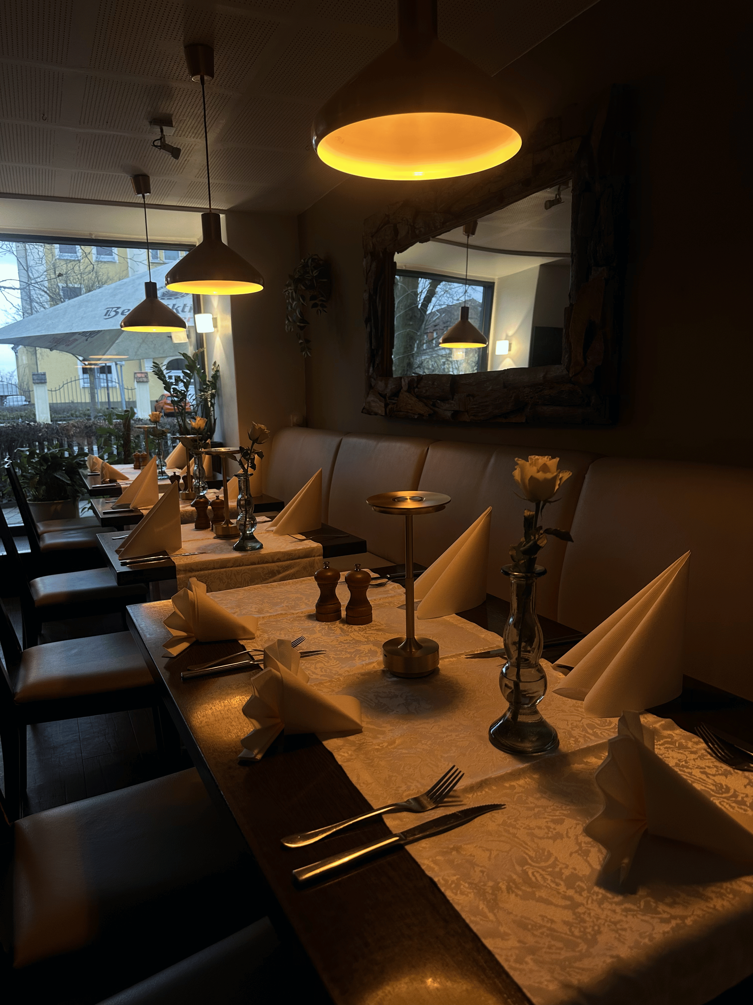 Ein gemütlich gedeckter Esstisch in einem Restaurant mit weißen Tischtüchern, Stoffservietten in Fächerform, Kerzenhaltern, Blumen und Gläsern, mit einer großen Fensterfront im Hintergrund, die einen Blick auf eine Außenfläche mit Sonnenschirm und Gebäuden bietet. Deckenleuchten sorgen für warmes Licht.