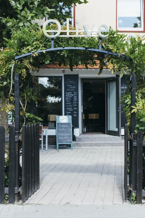 Eingang zu einem Restaurant namens 'Olive', umgeben von grünen Pflanzen, mit einem schwarzen Tor und einem grünen Schild, das die Speisen und Preise auflistet.