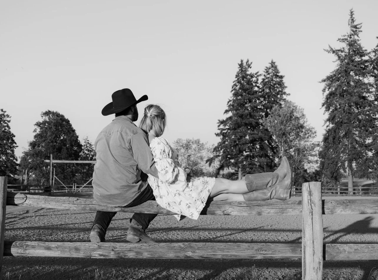 A man and woman sitting on a wooden fence outdoors, with tall trees in the background. The man is dressed in cowboy attire with a wide-brimmed hat, and the woman is wearing a floral dress with boots. They are facing away from the camera.