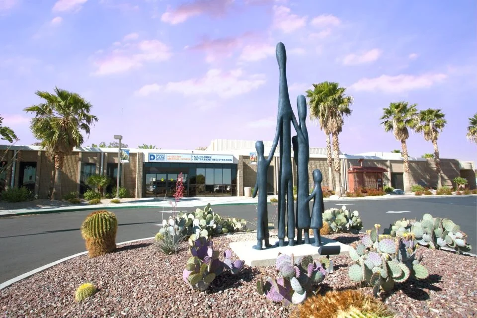 A sculpture of a family with five figures, in an outdoor desert landscape with cacti and palm trees, in front of a building with a sign that reads 'Main Lobby' and 'Outpatient Registration'.