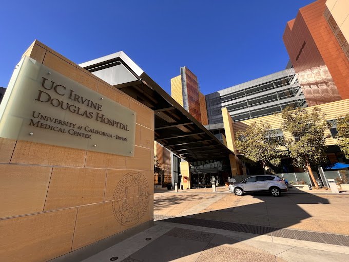 Exterior view of UC Irvine Douglas Hospital, part of the University of California Irvine Medical Center, with a parking lot and modern buildings.