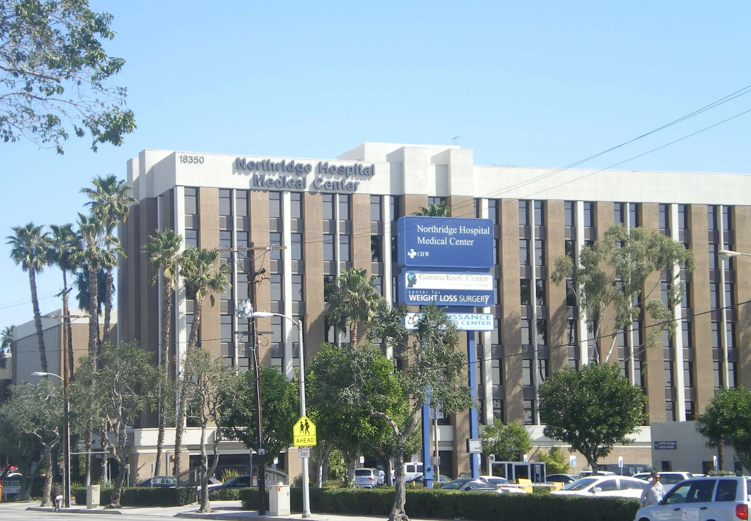 A multi-story hospital building with signs indicating it is Northridge Hospital Medical Center and associated health care services, with trees, cars, and pedestrians outside.