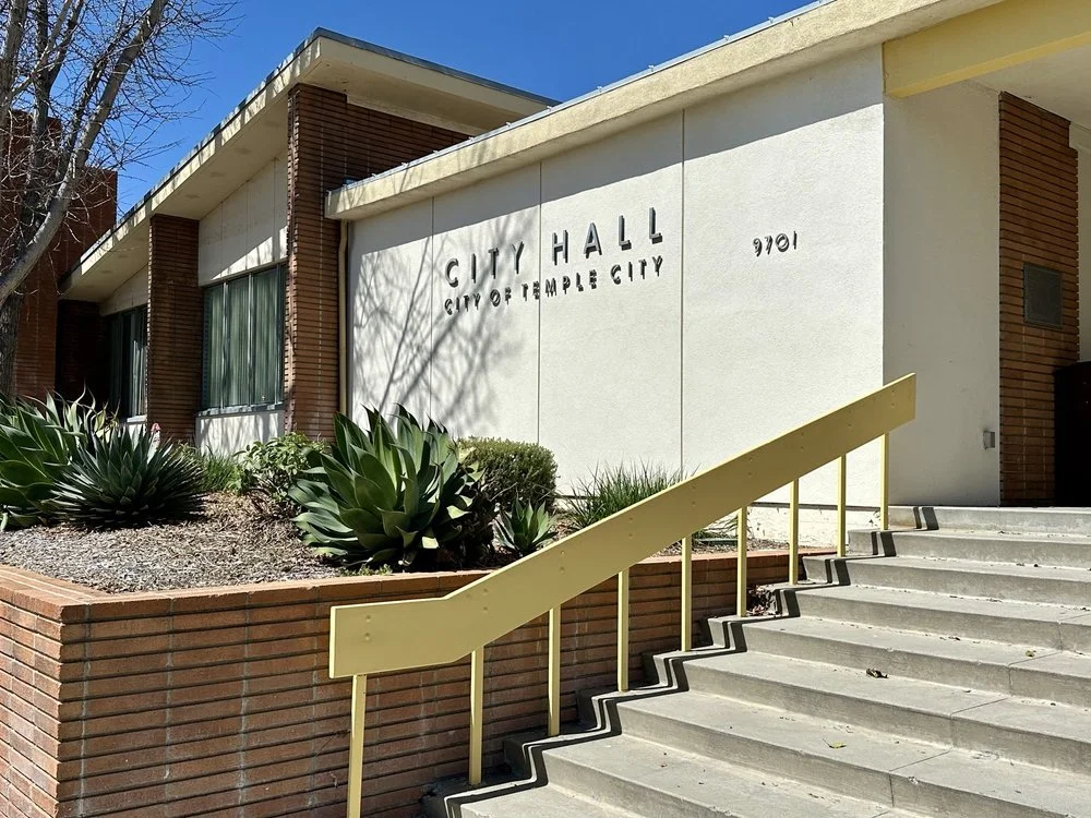 Exterior view of a city hall building with white walls, brick accents, and a yellow handrail along concrete stairs. There are large succulent plants in a raised brick planter in front. The building has signs indicating it is the City Hall of Temple C