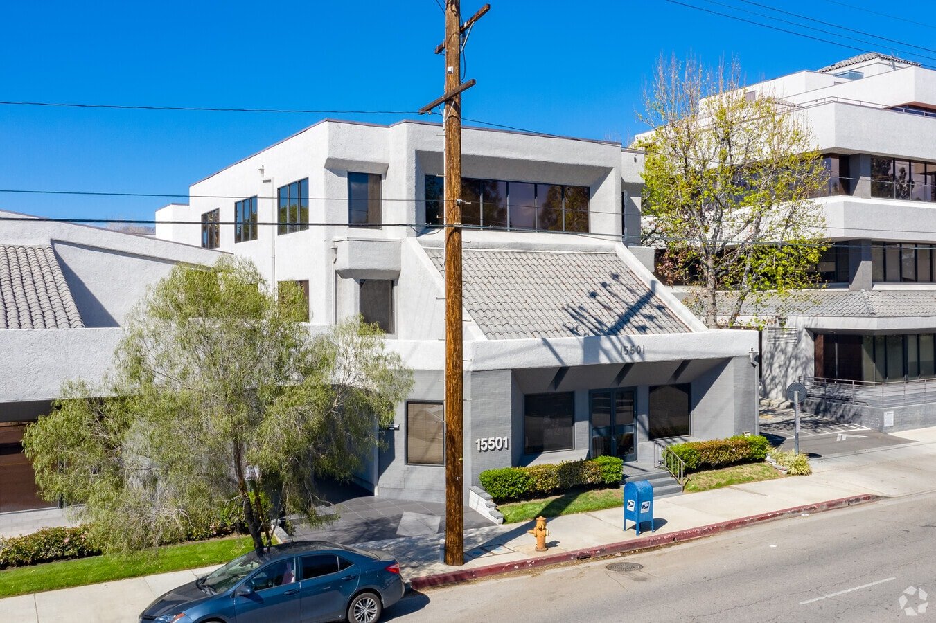 Modern white multi-story apartment building on city street with trees, a blue mailbox, a car, utility pole, and power lines.