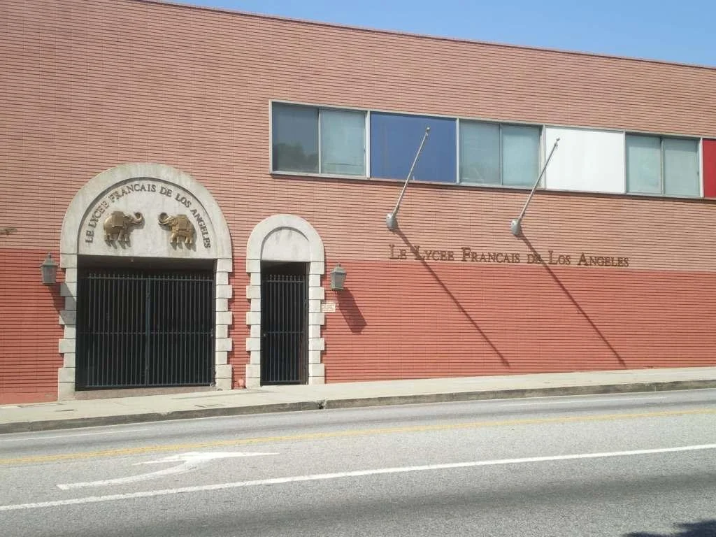 The exterior of a school named "Lycée Français de Los Angeles" with two adjacent gates and decorative elephant sculptures above the entrance, on a street with a clear sky.
