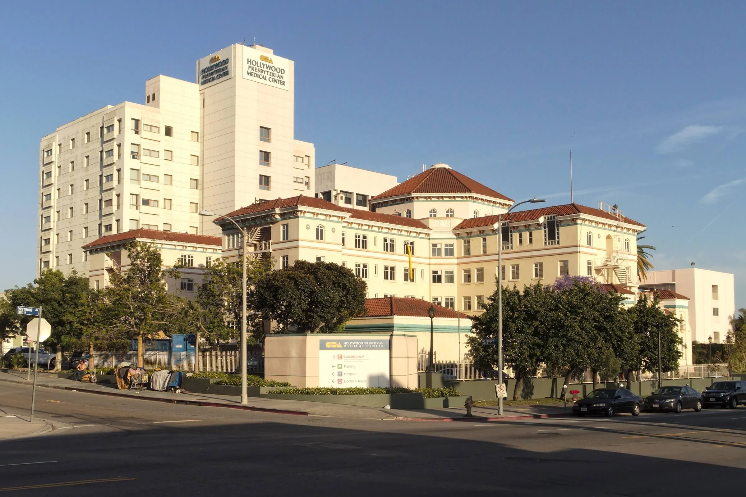 A large, white hospital building with a red-tiled roof, trees in front, on a sunny day, with a street and cars in the foreground.