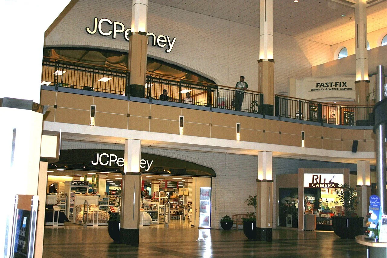 Inside a shopping mall featuring a JCPenney store on the ground and upper levels, with a sign for FAST-FIX jewelry and watch repairs on the upper level. There are potted plants and a camera shop sign near the entrance.