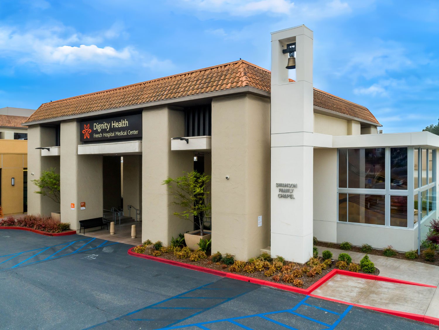 Exterior view of Dignity Health hospital building with a sign showing 'Dignity Health French Hospital Medical Center' and 'Swanson Family Chapel', parking spaces, landscaping, and a blue sky with clouds.