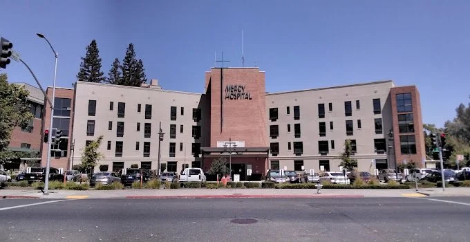 Mercy Hospital building with a parking lot in front, trees, and clear blue sky.