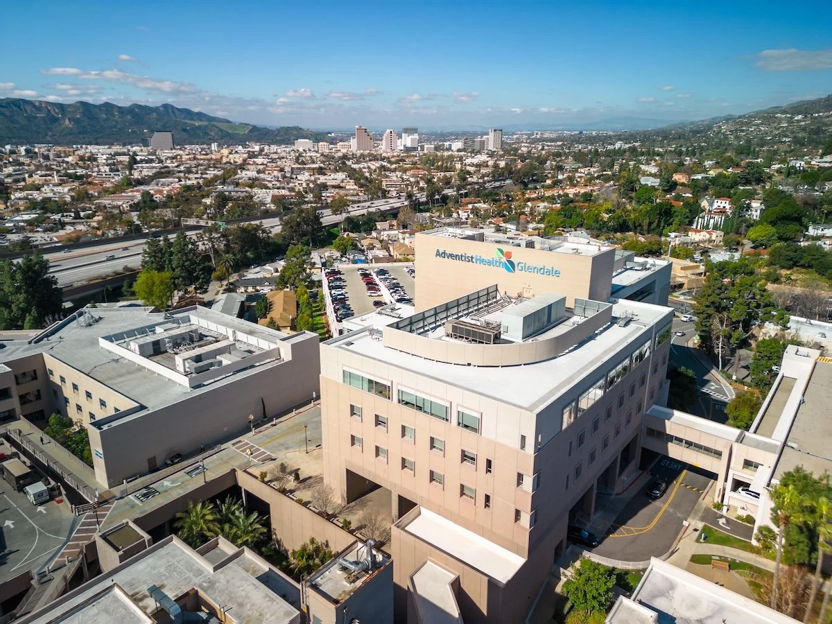 Aerial view of a hospital building with the sign 'Adventist Health Glendale,' surrounded by parking lots and cityscape with mountains in the background.