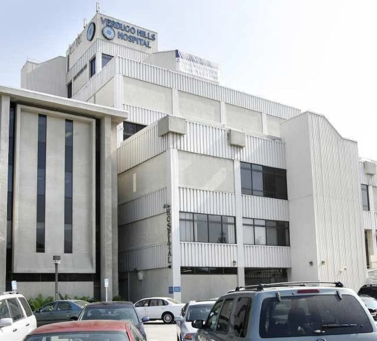Hospital building with a sign that says 'Venoco Hills Hospital' at the top, multiple parked cars in the foreground.