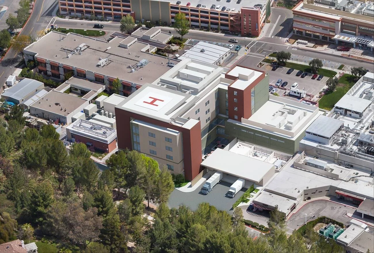 Aerial view of a medical building complex with a prominent blue 'H' on the roof, surrounded by parking lots, greenery, and other commercial buildings.