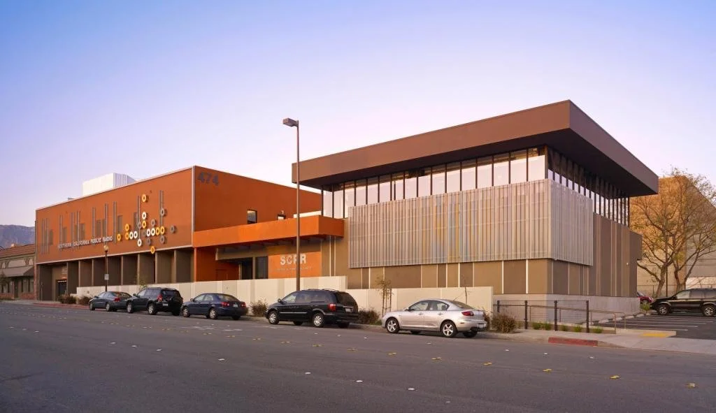 Modern architectural building with orange and gray exterior, parked cars along the street, and a lamppost in foreground.