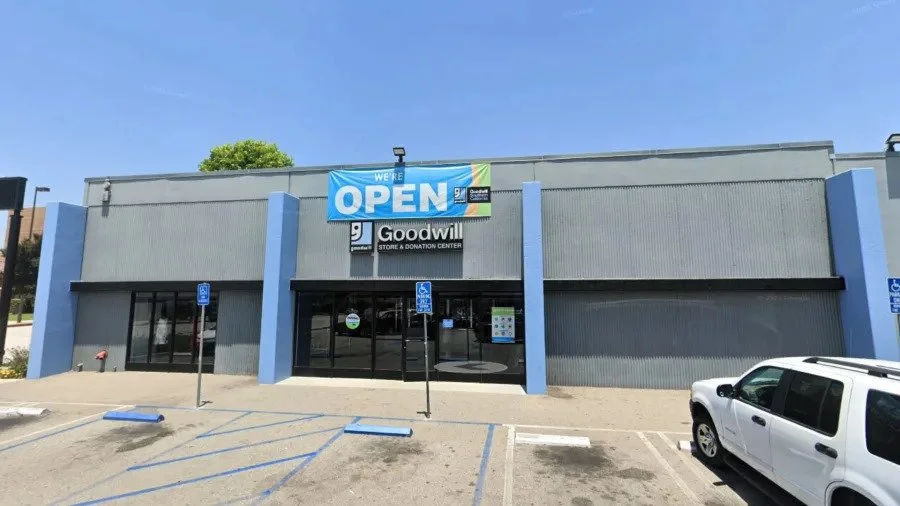 Front of a Goodwill store with a large blue and green open sign, parking spaces including handicapped spots, and a white vehicle parked outside.