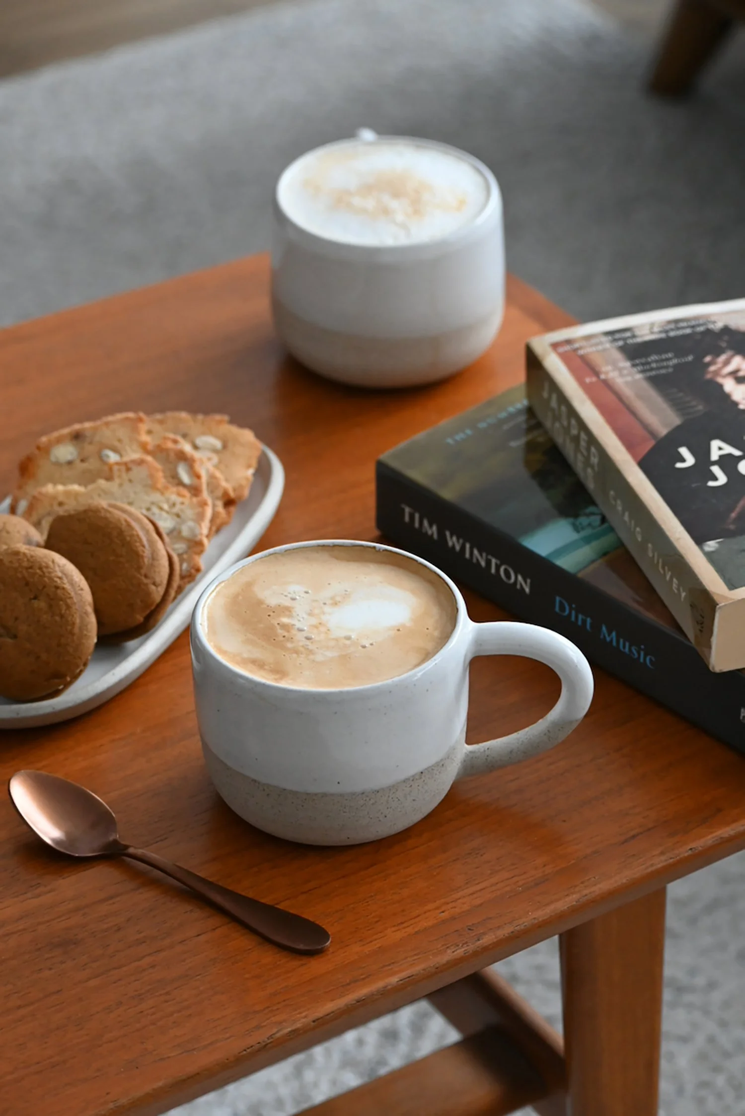 A wooden table with a white ceramic mug of frothy cappuccino, a spoon, a tray of cookies, and two books.