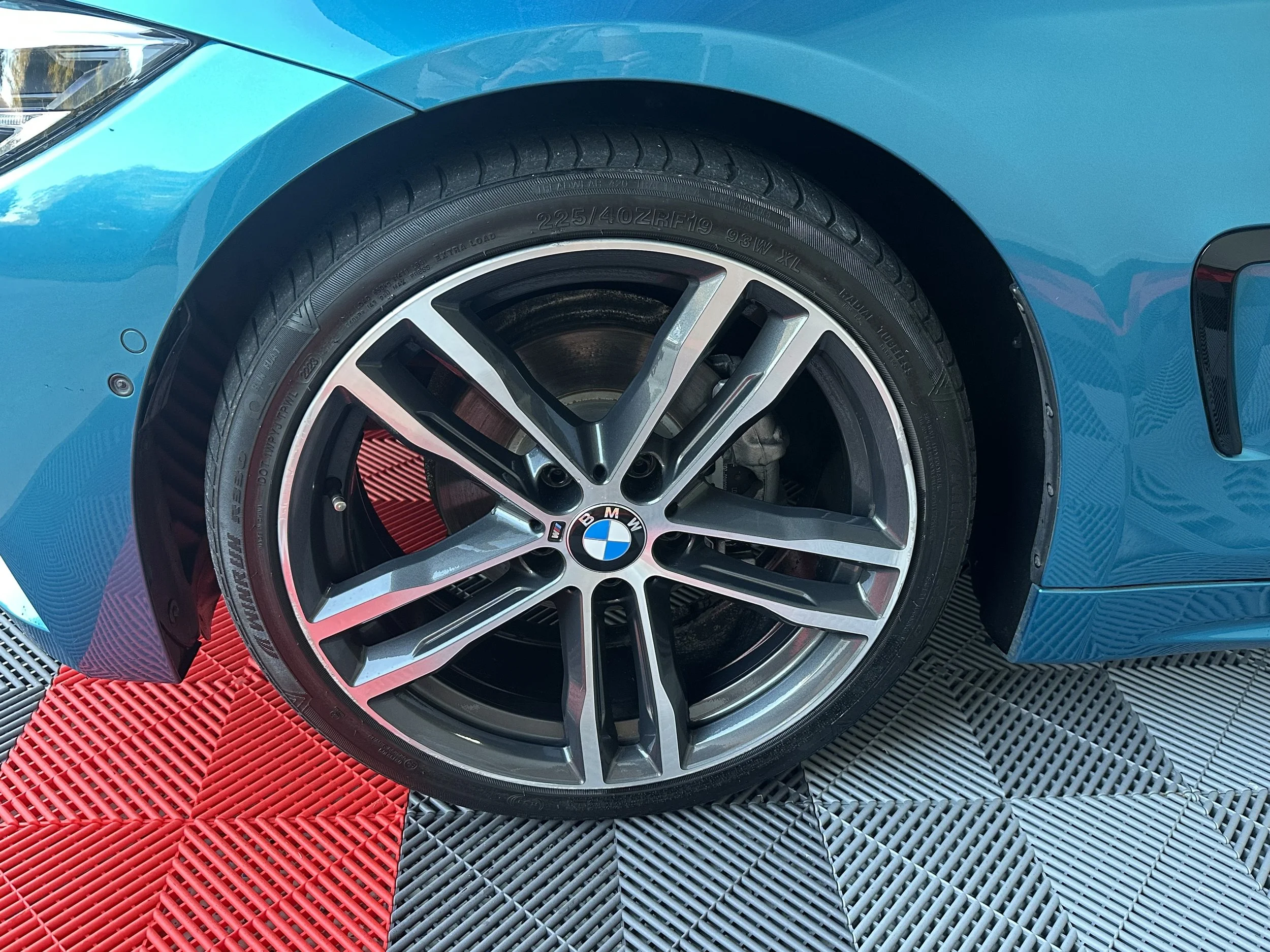 Close-up of a blue BMW car's front wheel, alloy rim, and tire, on a red and black rubber mat.