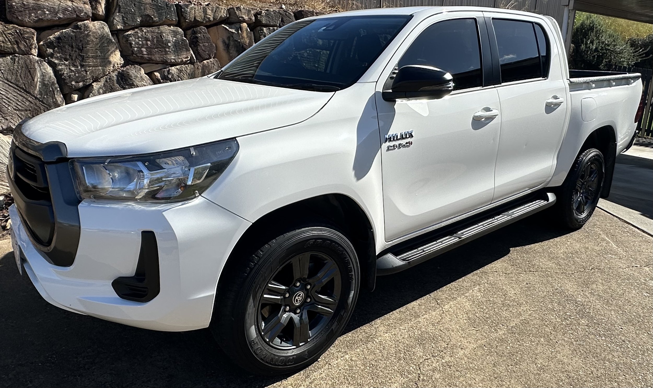 White Toyota pickup truck parked on a driveway with a stone wall and trees in the background.