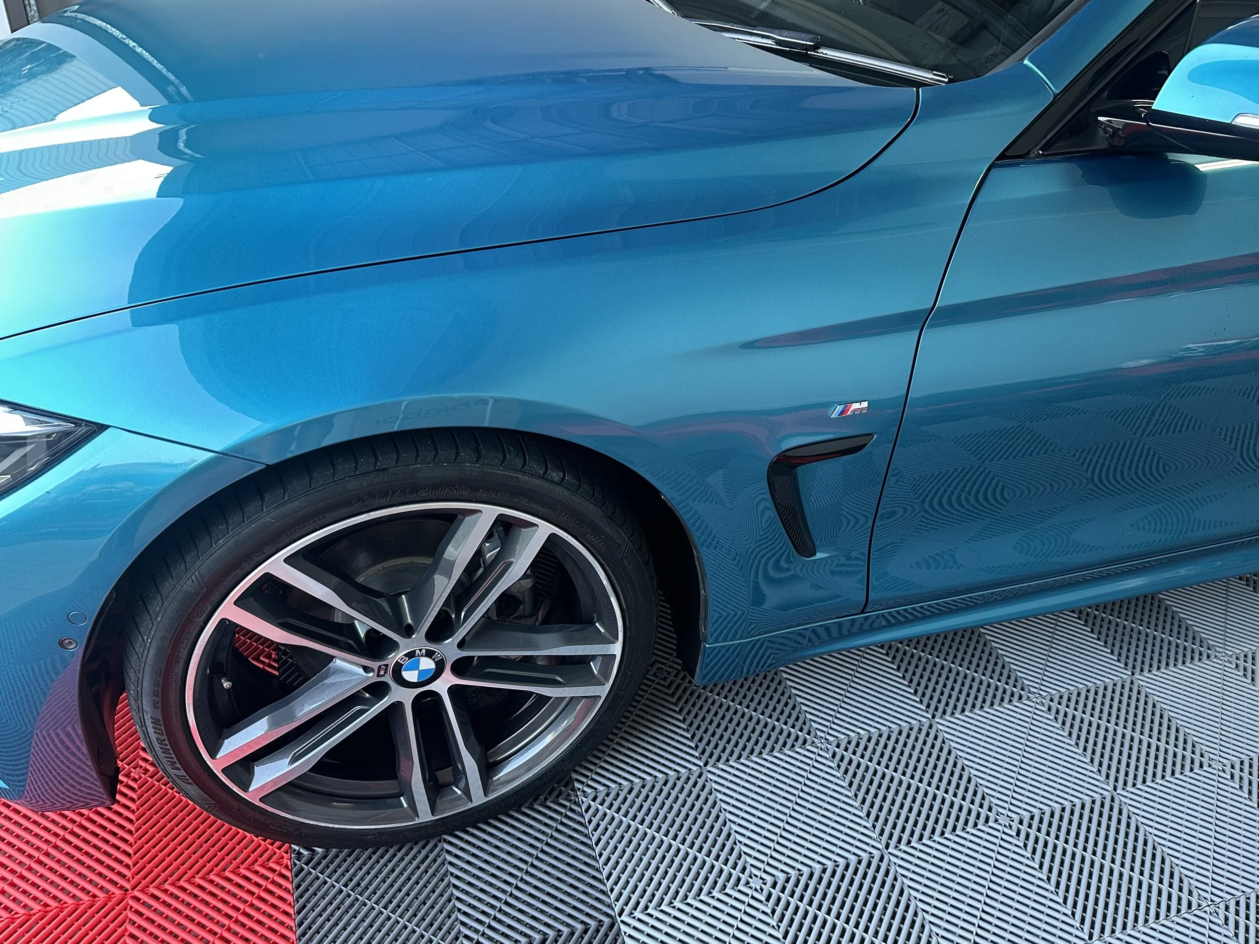 Close-up of a blue BMW M series car with a detailed wheel and BMW logo, parked on a black and red textured floor.