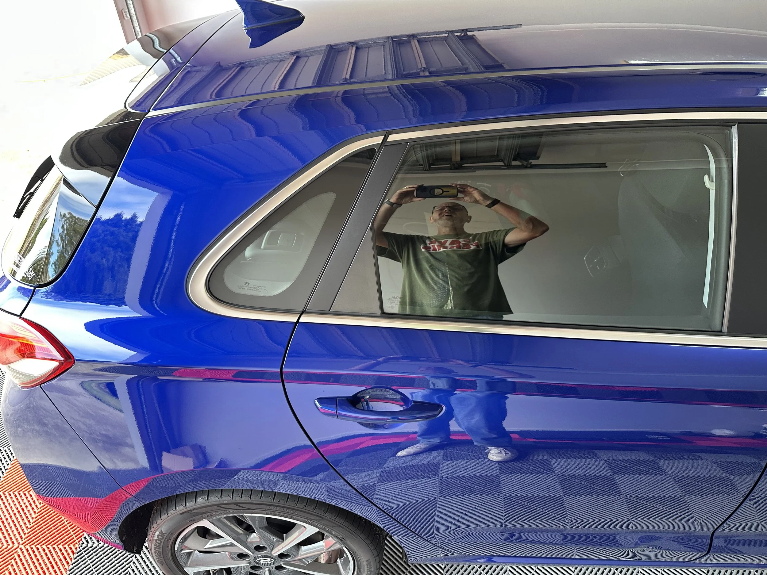 A person wearing glasses and a green t-shirt holding a camera takes a selfie in the reflection of a shiny blue car's side window, parked indoors.