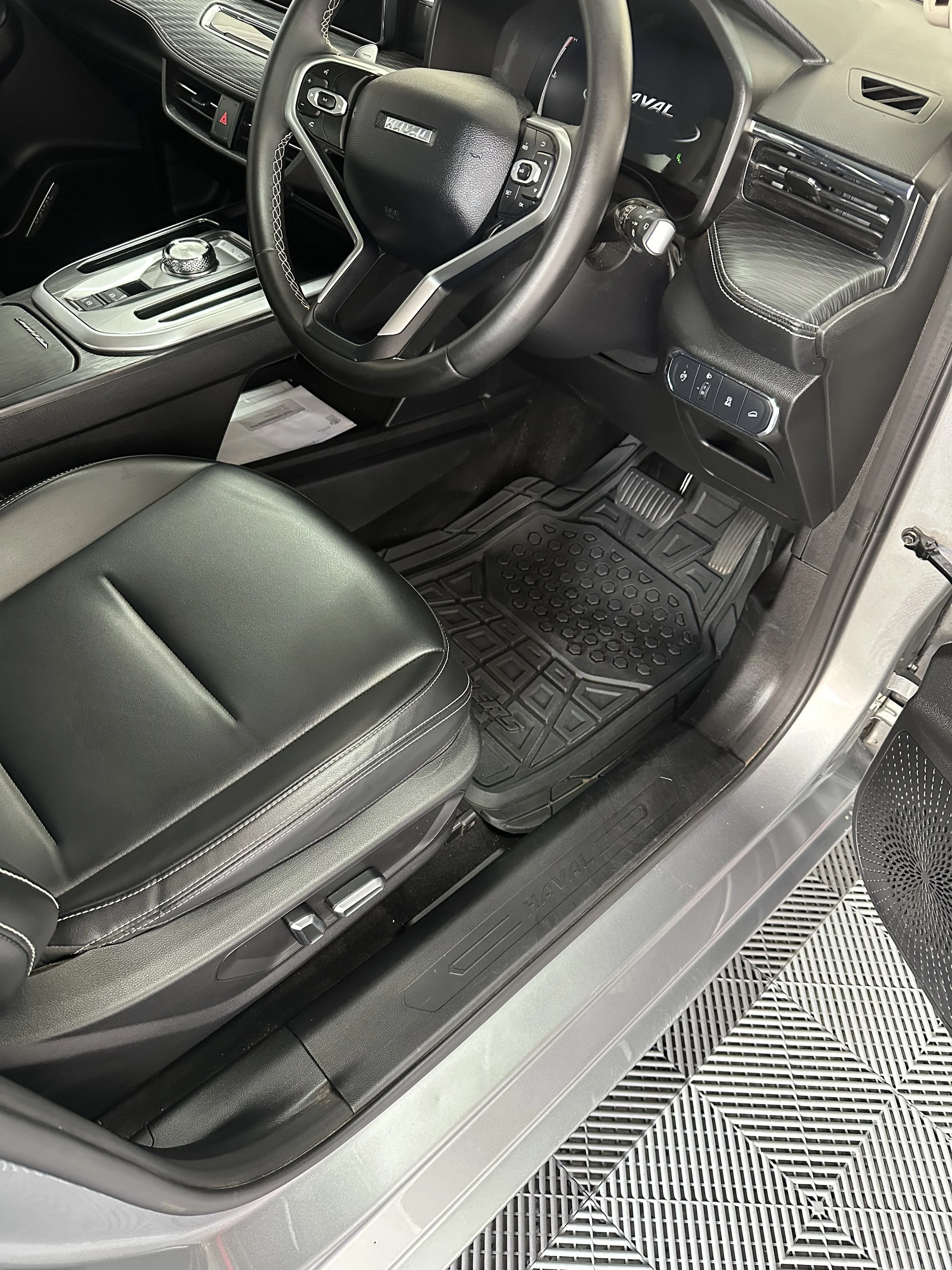 Interior view of a Land Rover vehicle showing the steering wheel, dashboard, center console, and driver's side floor mat with the Land Rover logo. The car appears to be parked on a metal grating with a textured floor mat.