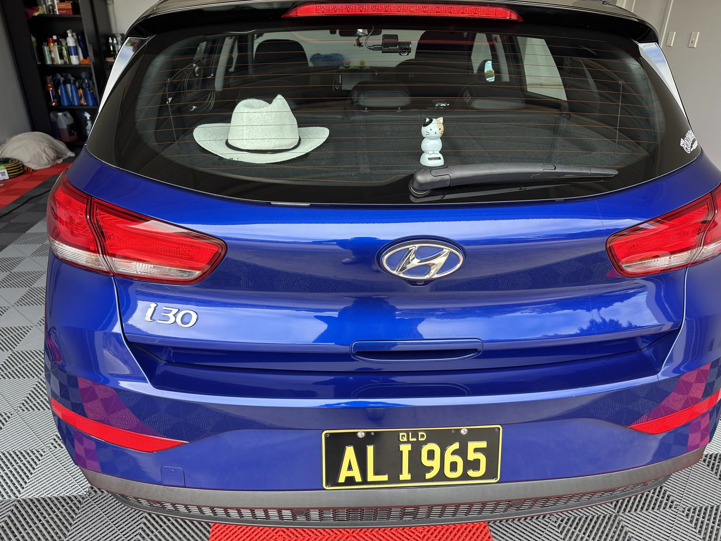 The rear view of a blue Hyundai I30 with a yellow license plate, a white hat, and a cat-shaped air freshener hanging from the rearview mirror, parked inside a garage with shelves and tools.