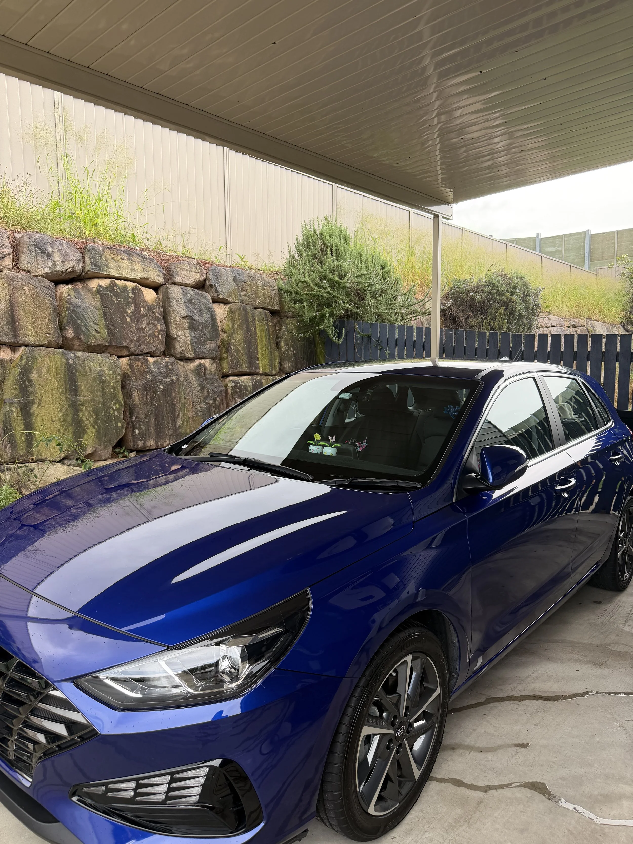 A blue modern hatchback car parked in a garage with a concrete floor, a stone retaining wall with greens and shrubs, and a beige fence and metal roof overhead.