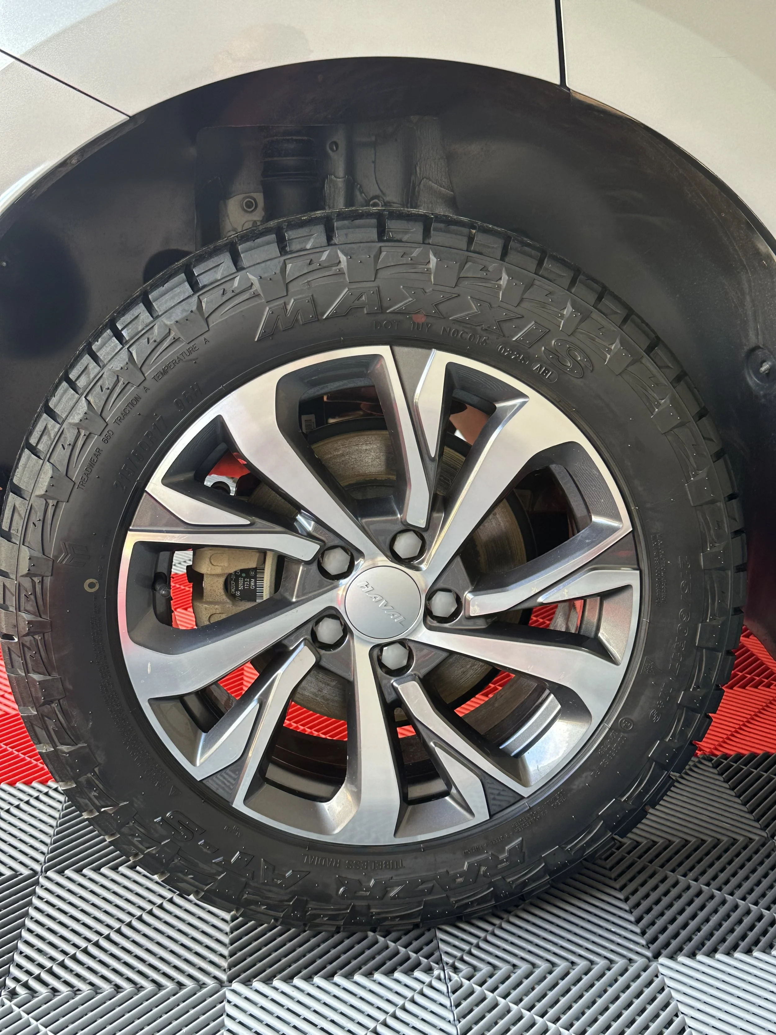 Close-up of a vehicle wheel featuring a Maxxis tire with a detailed tread pattern and an alloy rim, set on a textured metal surface.