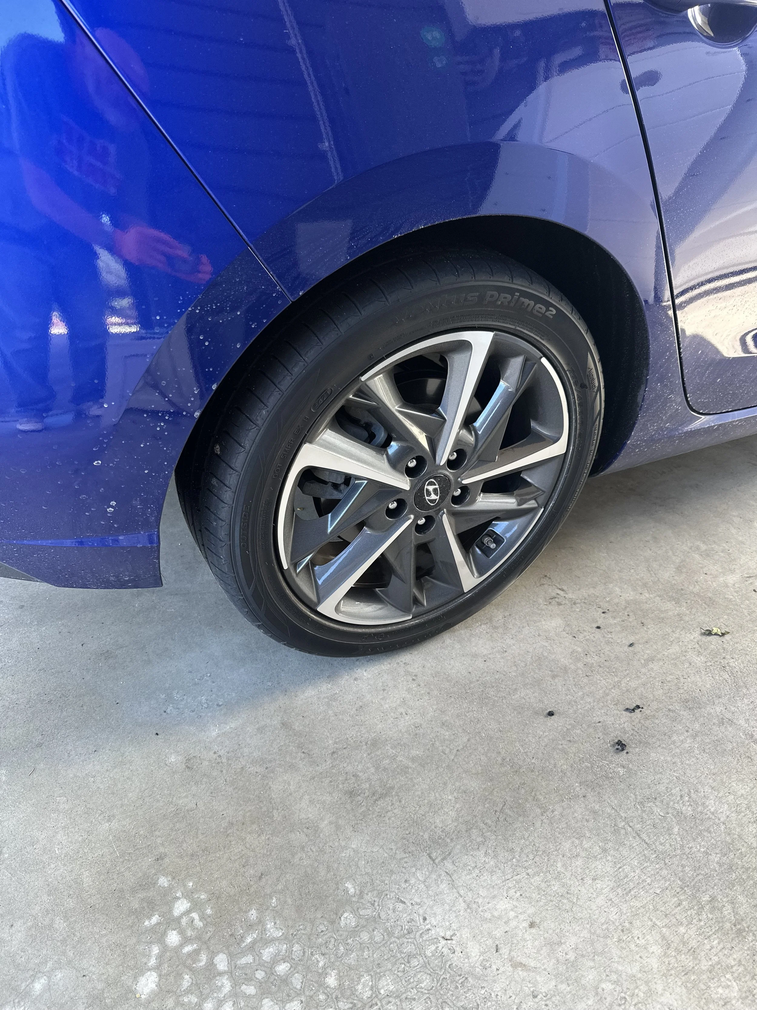 Close-up of a blue car showing the front wheel, part of the fender, and the reflection on the car's surface. The car is parked on a concrete surface.