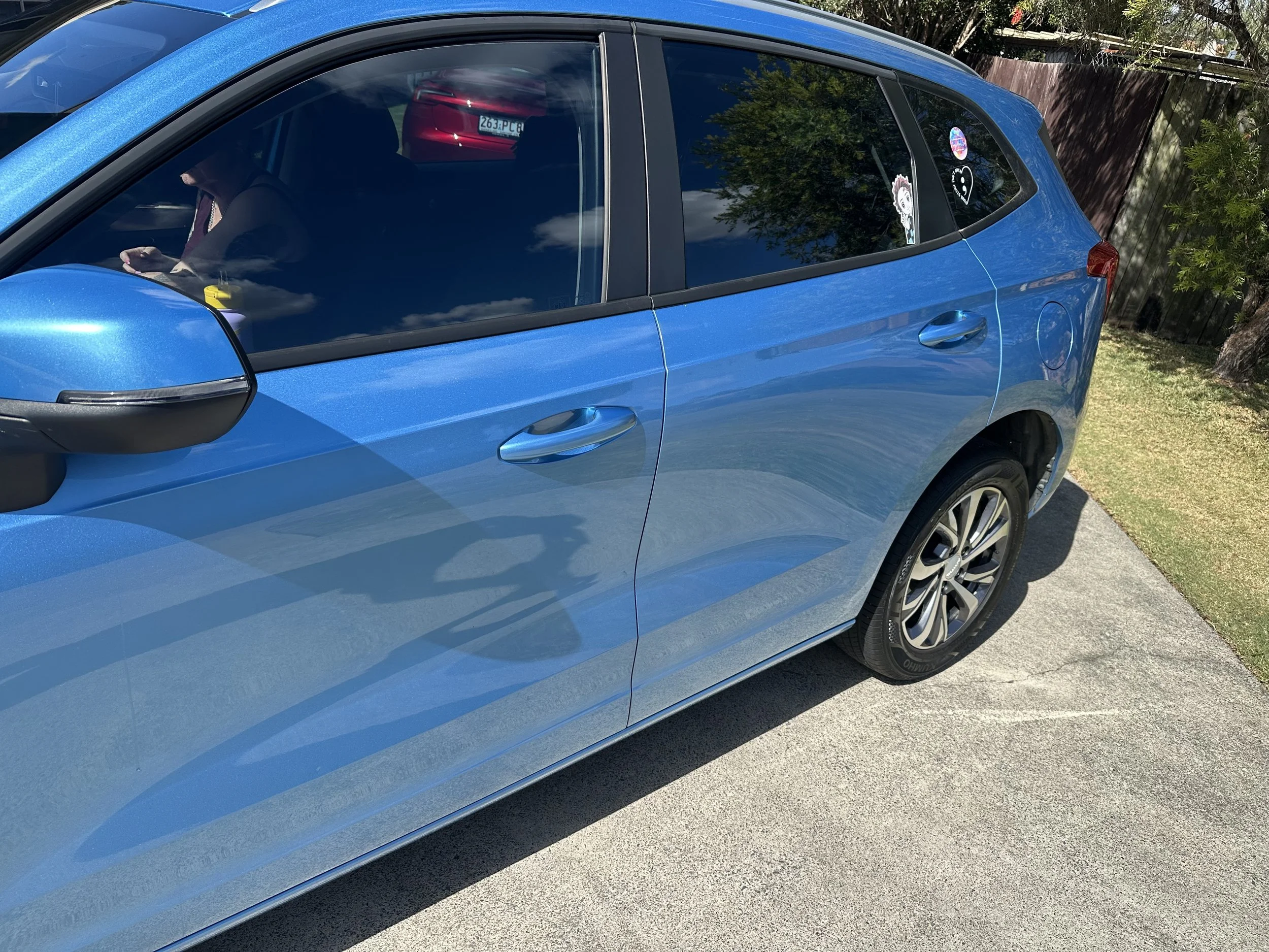 A blue hatchback car parked outdoors on a driveway, with a woman sitting inside looking at her phone, and stickers on the rear window including a cartoon character and a heart-shaped design.