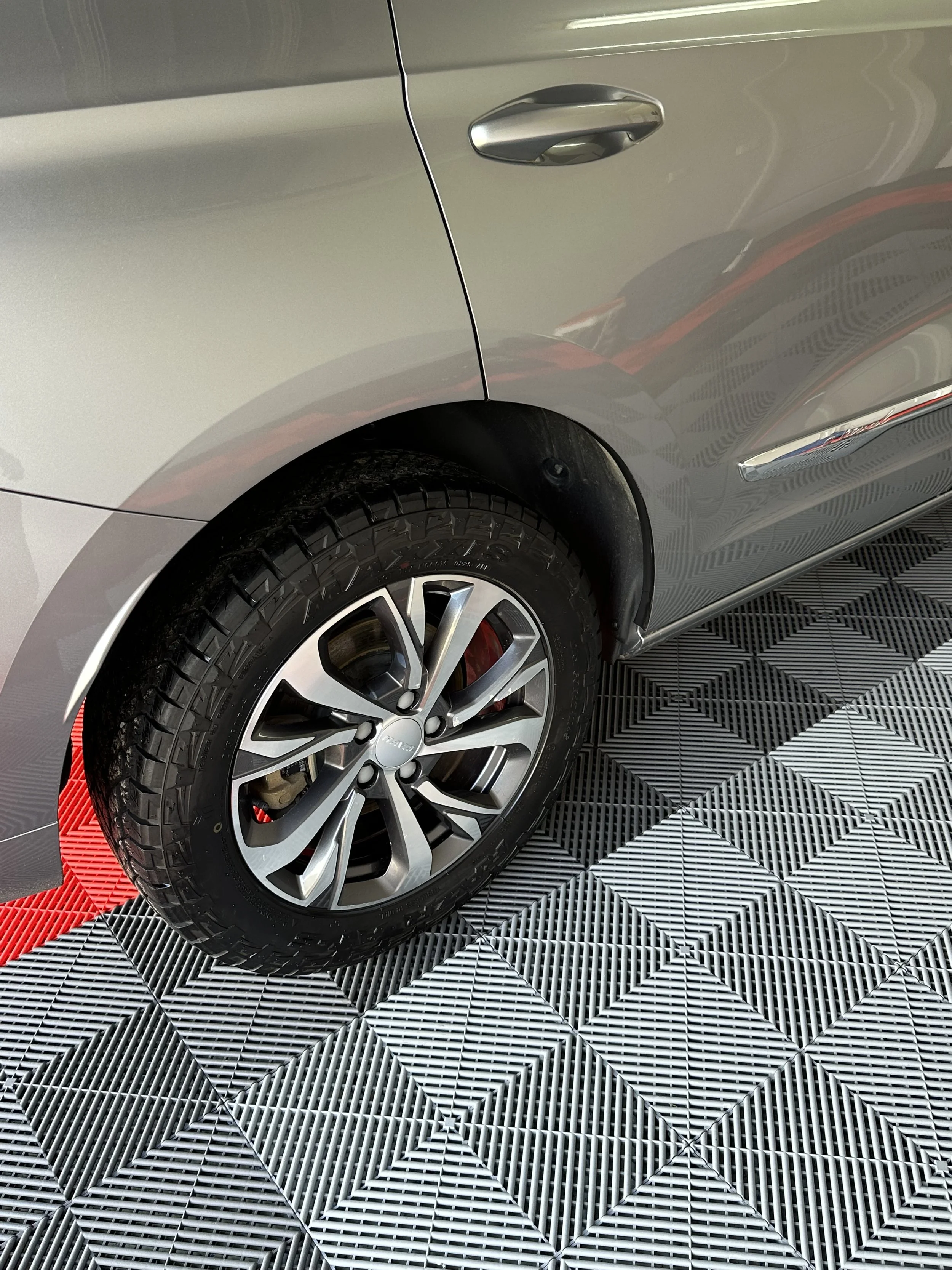 Close-up of a part of a silver car, focusing on the front right wheel and a portion of the front door and side panel, parked on a black and white geometric patterned floor.