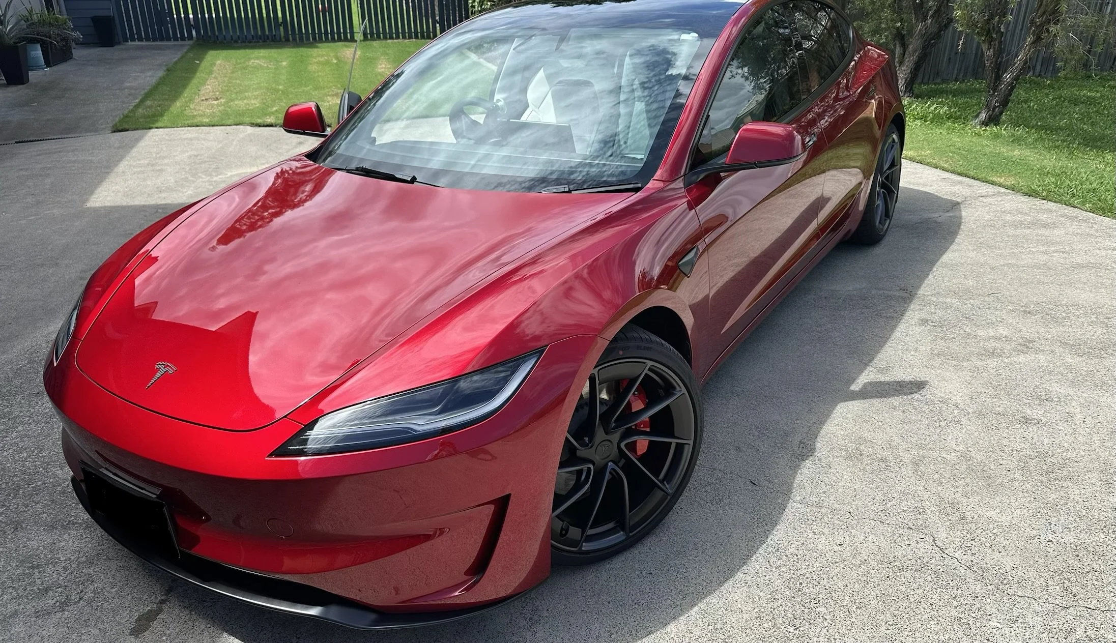 Red Tesla Model 3 parked on driveway next to grass and trees.